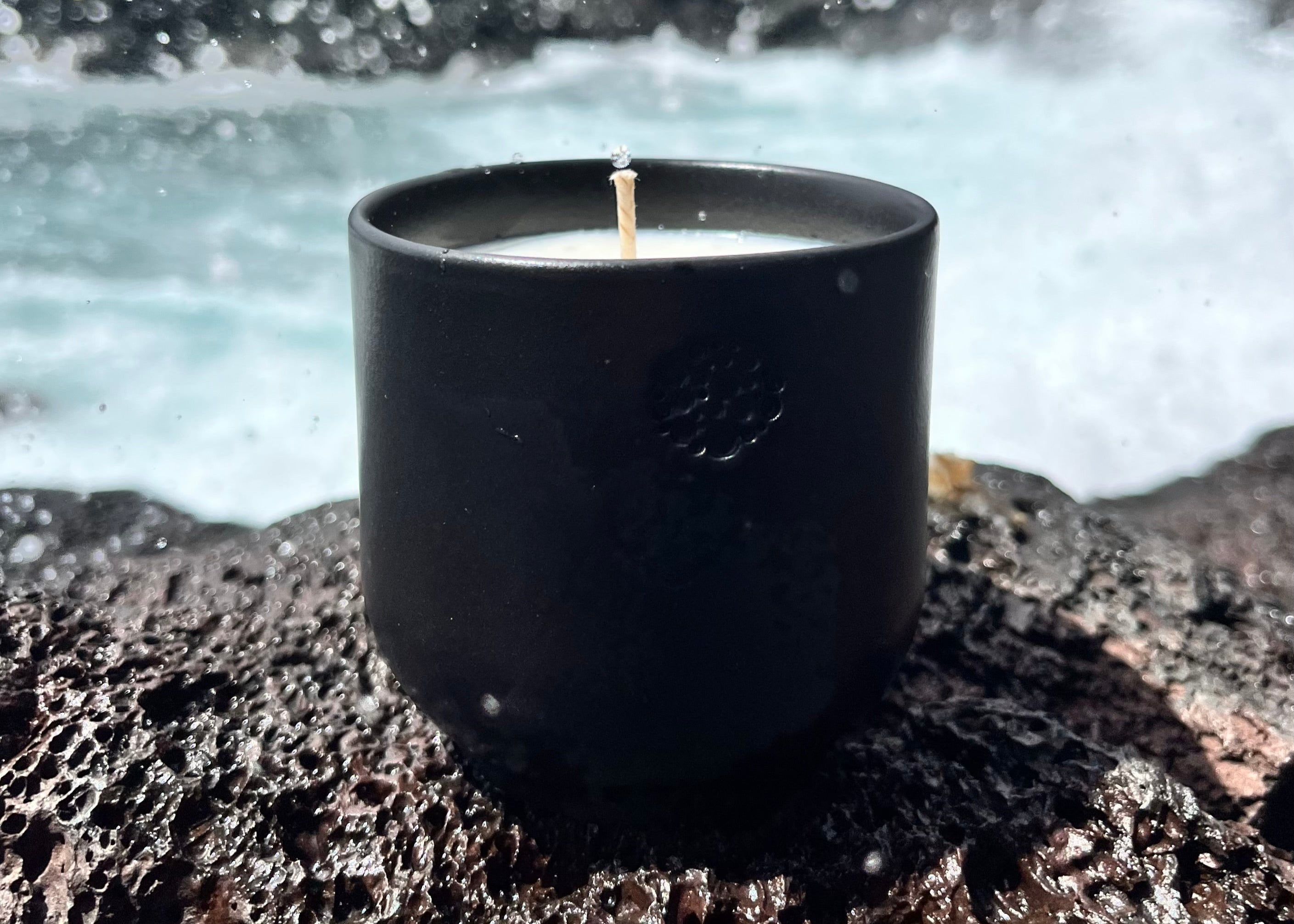 Eine schwarze Kerze steht auf einem Felsen aus Basalt, der besonders großporig und rau aussieht. Im Hintergrund sind die Wellen des Meeres verschwommen zu sehen. Die Spritzer einer Welle sind über das ganze Bild verteilt. 