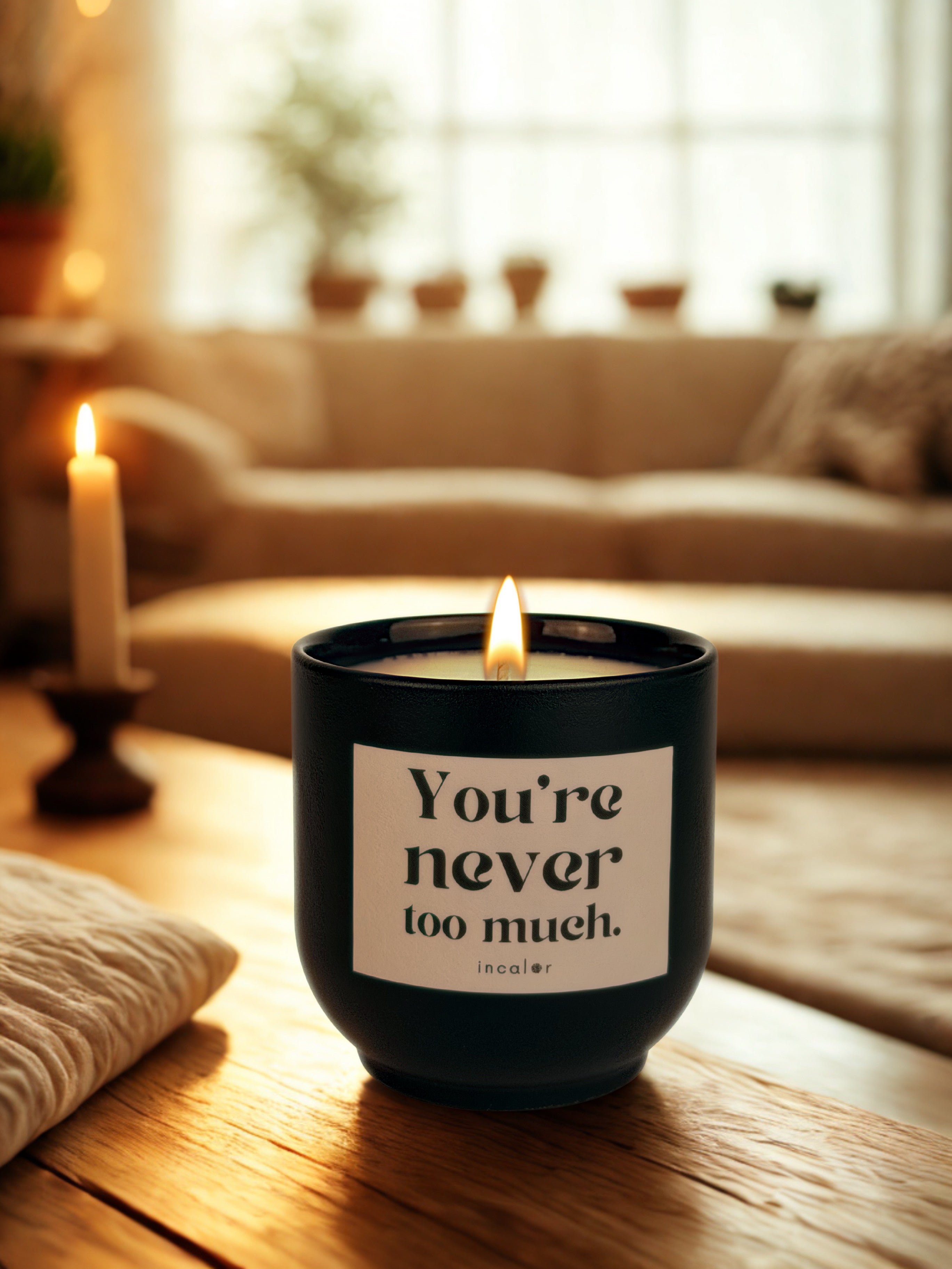 Dunkelgrüne (deep ivy) Duftkerze mit cremefarbenem Etikett und der Aufschrift „You're never too much.“ steht auf einem Holztisch in einem warm beleuchteten Wohnzimmer mit Sofa, Pflanzen und weiterer Kerzendeko im Hintergrund.
