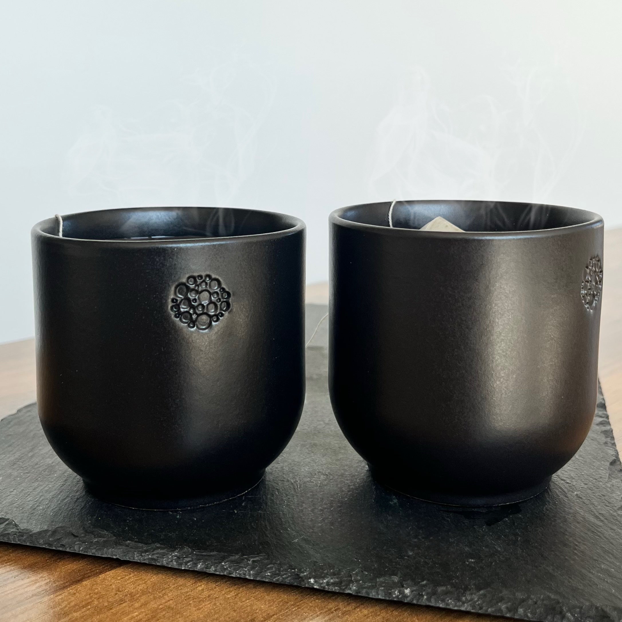 Zwei schwarze Incalor Becher stehen auf einer Schiefertafel, die wiederum auf einem hölzernen Untergrund liegt. Die Becher zeigen ihr geprägtes Incalor Logo, welches aus vielen kleinen Punkten und Kreisen besteht. In den Bechern hängen zwei Teebeutel und es steigt Dampf auf. Der Hintergrund ist minimalistisch und weiß. 
