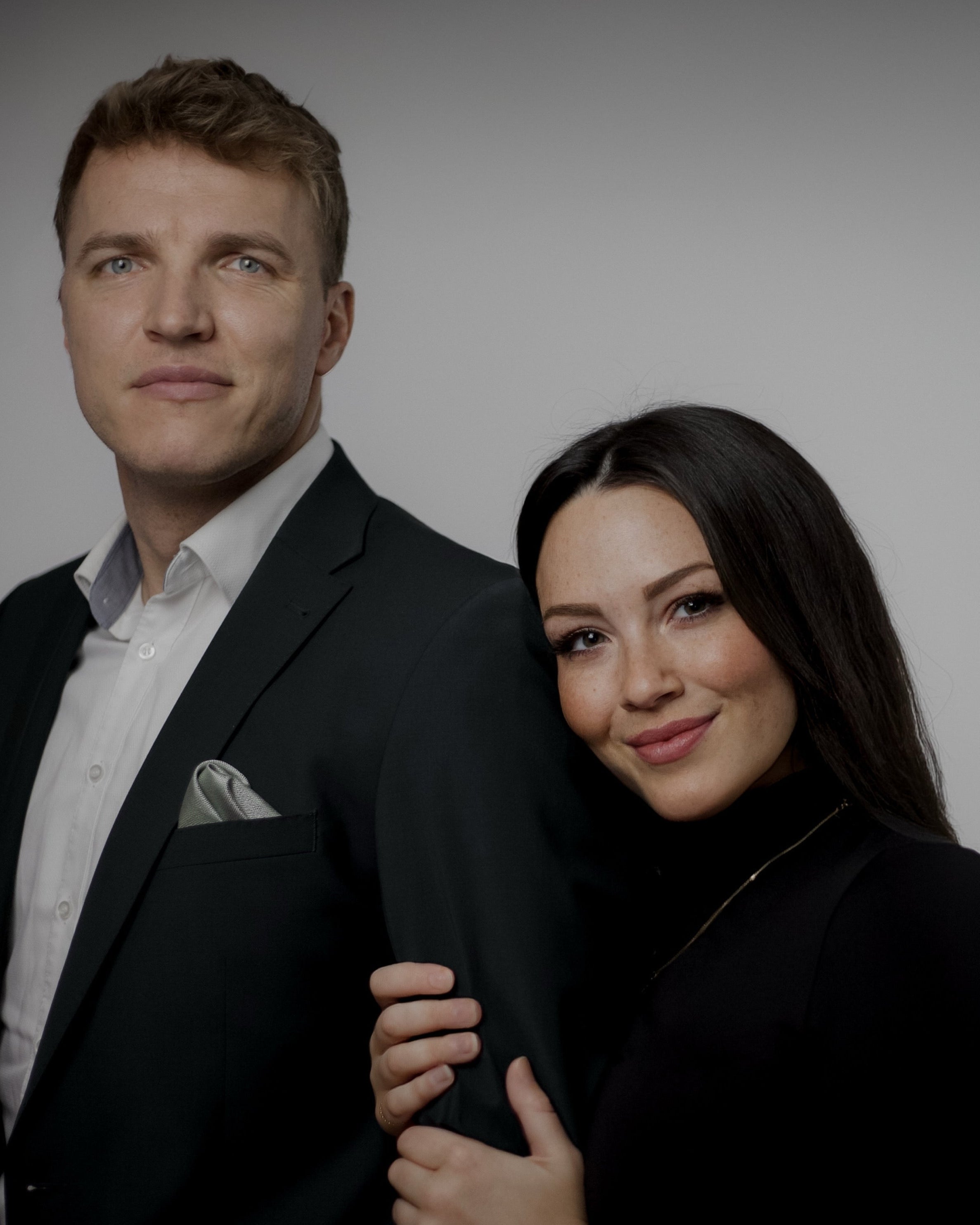 Professionelles Porträt von Matthias und Pauline vor grauem Hintergrund. Matthias trägt einen dunkelgrünen Anzug mit weißem Hemd und grauem Einstecktuch, hat kurze blonde Haare und blaue Augen. Pauline trägt einen schwarzen Rollkragenpullover und eine Armbanduhr, hat langes schwarzes Haar und lächelt warm in die Kamera. Sie steht leicht seitlich zu ihm und hält ihren Arm um seinen Arm. Das Foto ist im Studio-Stil aufgenommen und vermittelt eine formelle Atmosphäre.