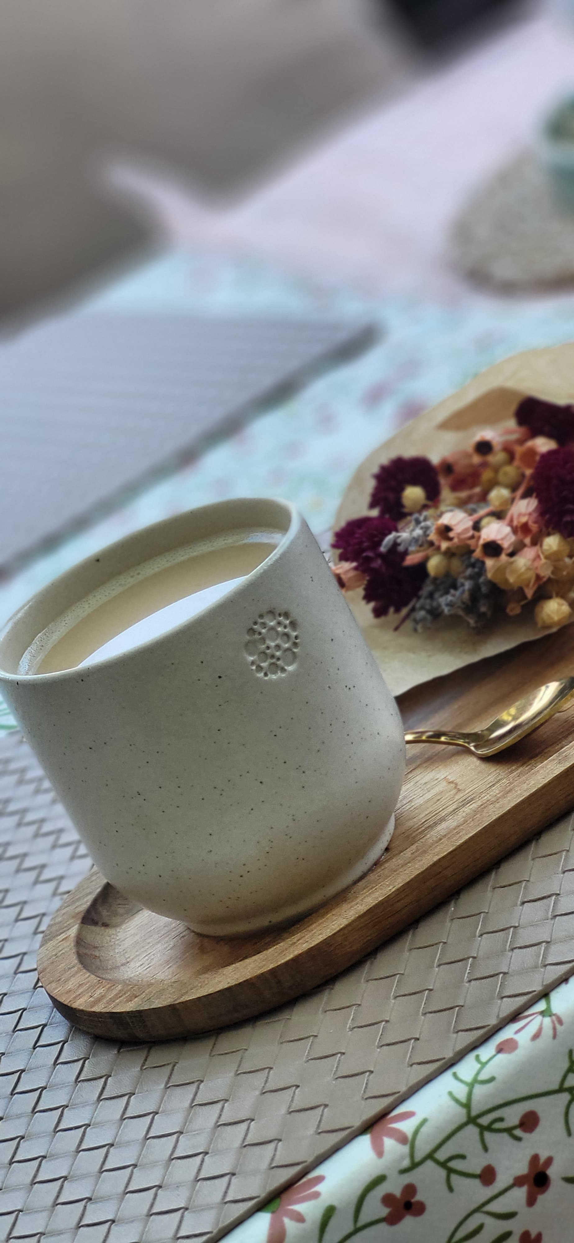 Eine Keramiktasse in sahara mit hellbraunem Kaffee oder Tee steht auf einem ovalen Holztablett. Daneben liegt ein kleiner Strauß getrockneter Blumen in verschiedenen Farben - dunkelrote, rosa, gelbe und blaue Blüten. Das Tablett steht auf einer geflochtenen Platzdecke mit Zickzackmuster. Im Hintergrund sind verschwommen weiße und hellblaue Gegenstände sowie Stoff mit kleinen Blumenmuster zu erkennen. Die Szene wirkt gemütlich und dekorativ arrangiert.