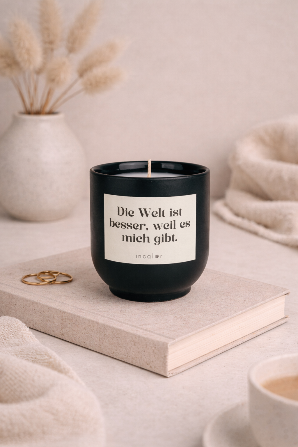 Dunkelgrüne (deep ivy) Kerzen-Tasse mit der Aufschrift ‚Die Welt ist besser, weil es mich gibt‘ steht auf einem beigen Buch. Daneben liegen goldene Ringe, im Hintergrund sorgen eine Vase mit Trockenblumen und weiche Textilien für eine ruhige, warme Wohlfühlatmosphäre.