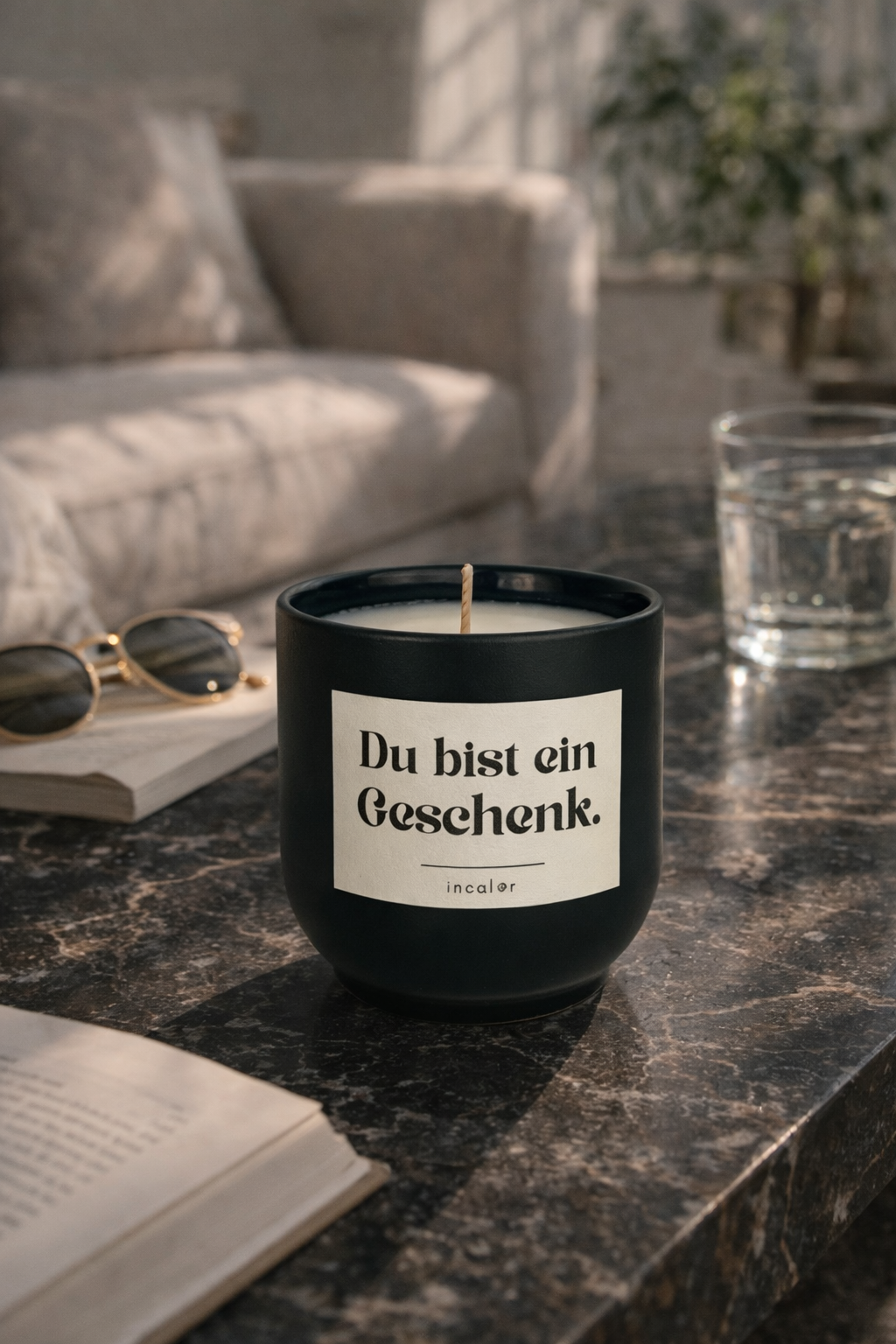 Dunkelgrüne (deep ivy) Duftkerze mit der Aufschrift ‚Du bist ein Geschenk‘ steht auf einem dunklen Marmortisch. Im Hintergrund befinden sich ein Sofa, eine Zimmerpflanze, ein Glas Wasser sowie eine Sonnenbrille und ein geöffnetes Buch, wodurch eine entspannte, wohnliche Atmosphäre entsteht.