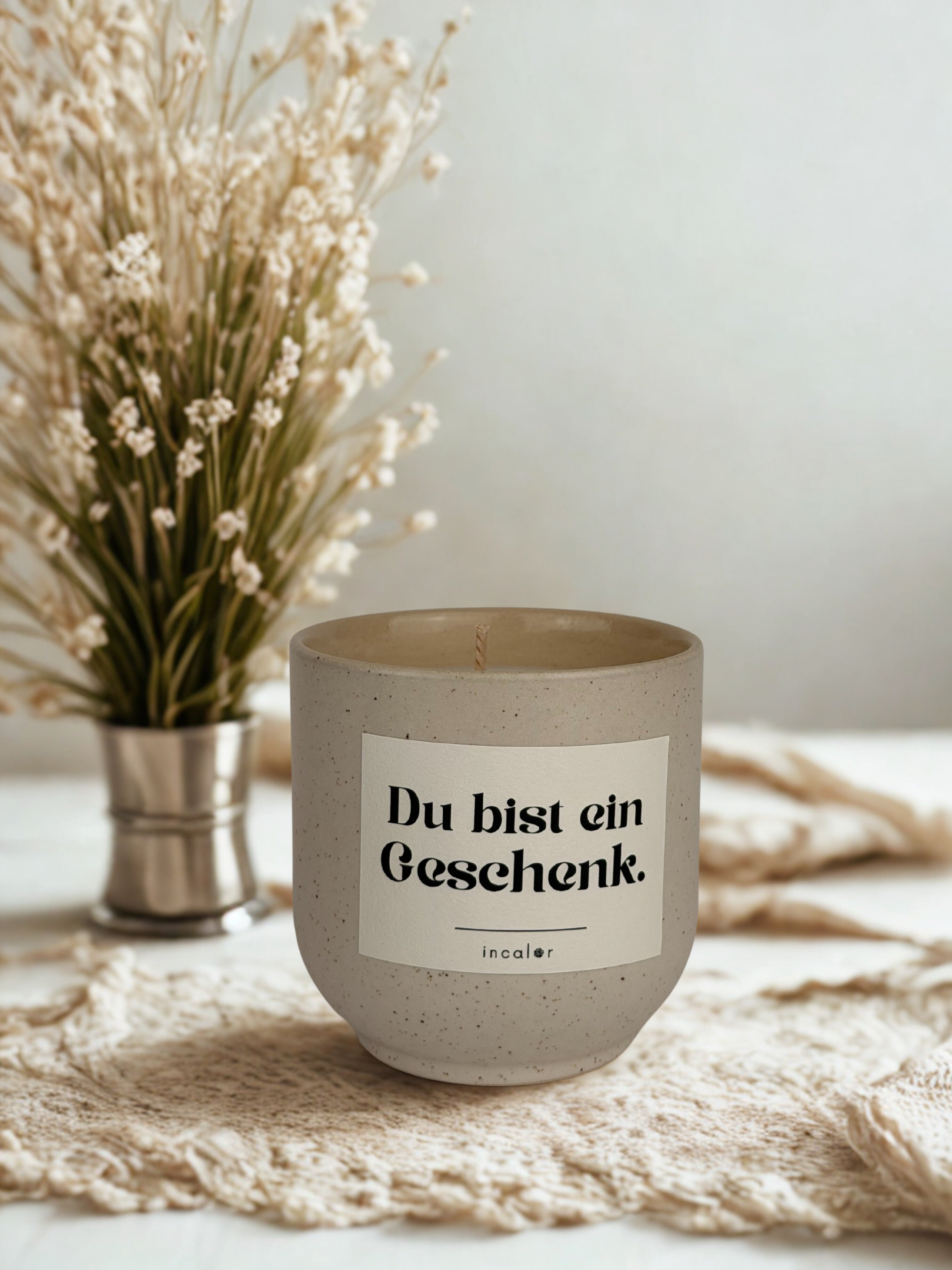 Beige (sahara), gesprenkelte Kerze mit der Aufschrift ‚Du bist ein Geschenk‘ steht auf einem hellen, strukturierten Stoff. Im Hintergrund ist ein kleiner Metallbecher mit getrockneten Blumen zu sehen, was eine ruhige, natürliche Atmosphäre schafft.