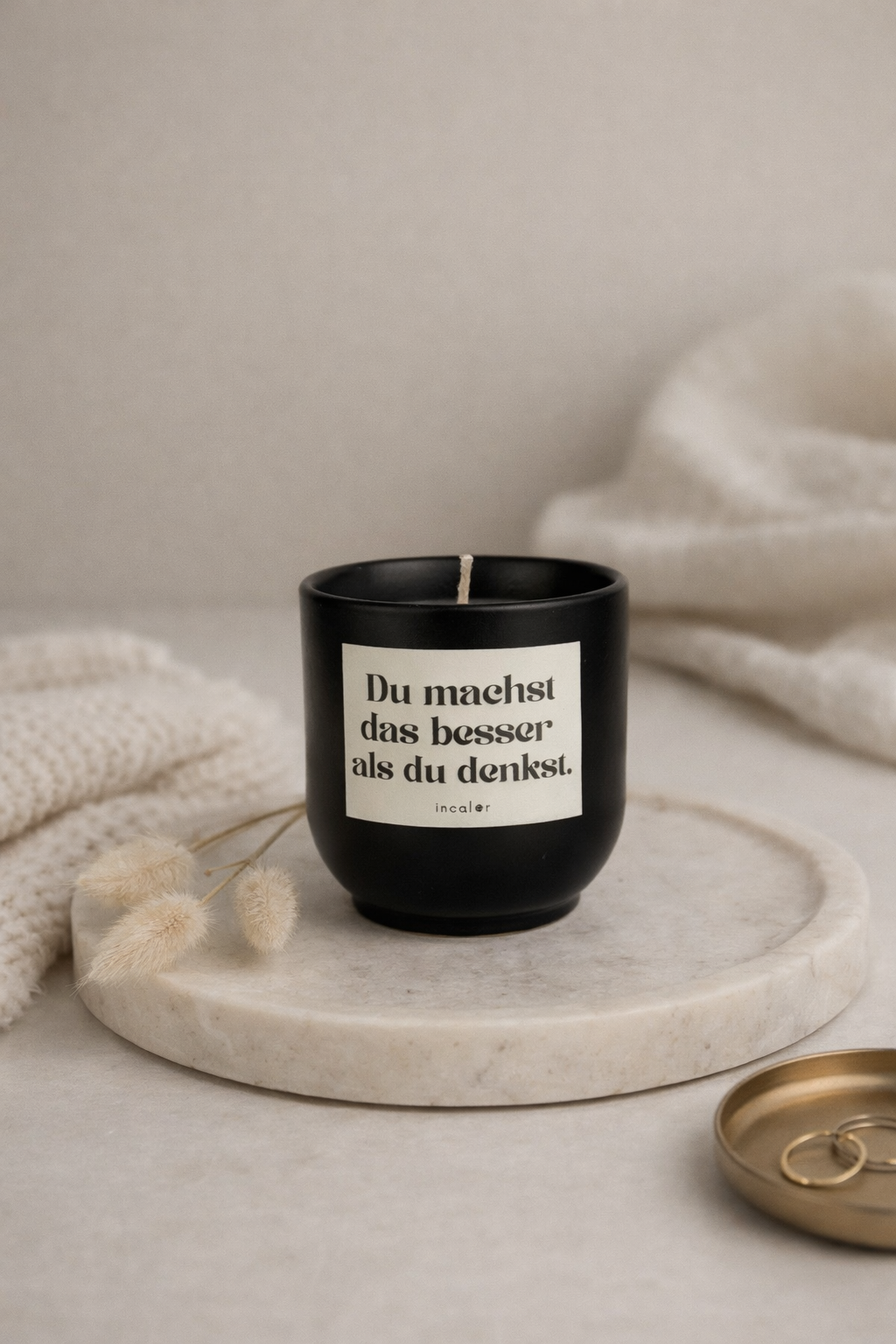 Schwarze (basalt) Kerze mit der Aufschrift ‚Du machst das besser als du denkst.‘ steht auf einem runden Marmortablett. Daneben liegen eine weiche Stoffdecke, getrocknete Gräser und eine kleine Schale mit Schmuck, wodurch eine ruhige, minimalistische Atmosphäre entsteht.