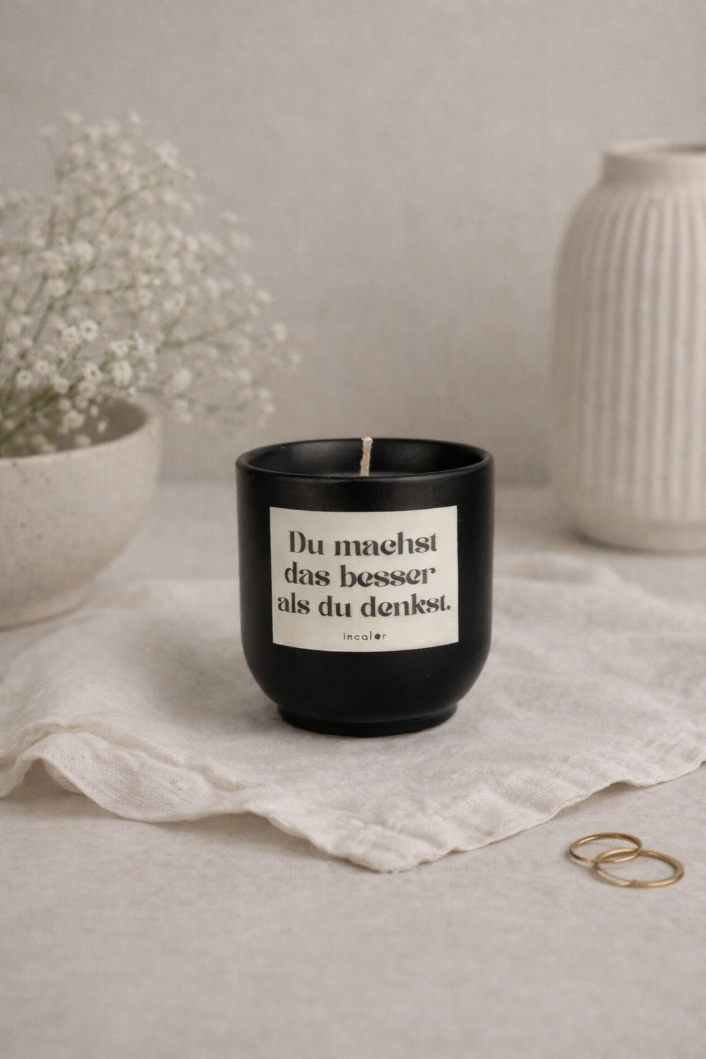 Schwarze (basalt) Kerze mit der Aufschrift ‚Du machst das besser als du denkst.‘ steht auf einem hellen Stofftuch. Daneben liegen zwei goldene Ringe sowie eine Vase und eine Schale mit feinen Trockenblumen, was eine ruhige, minimalistische Atmosphäre schafft.