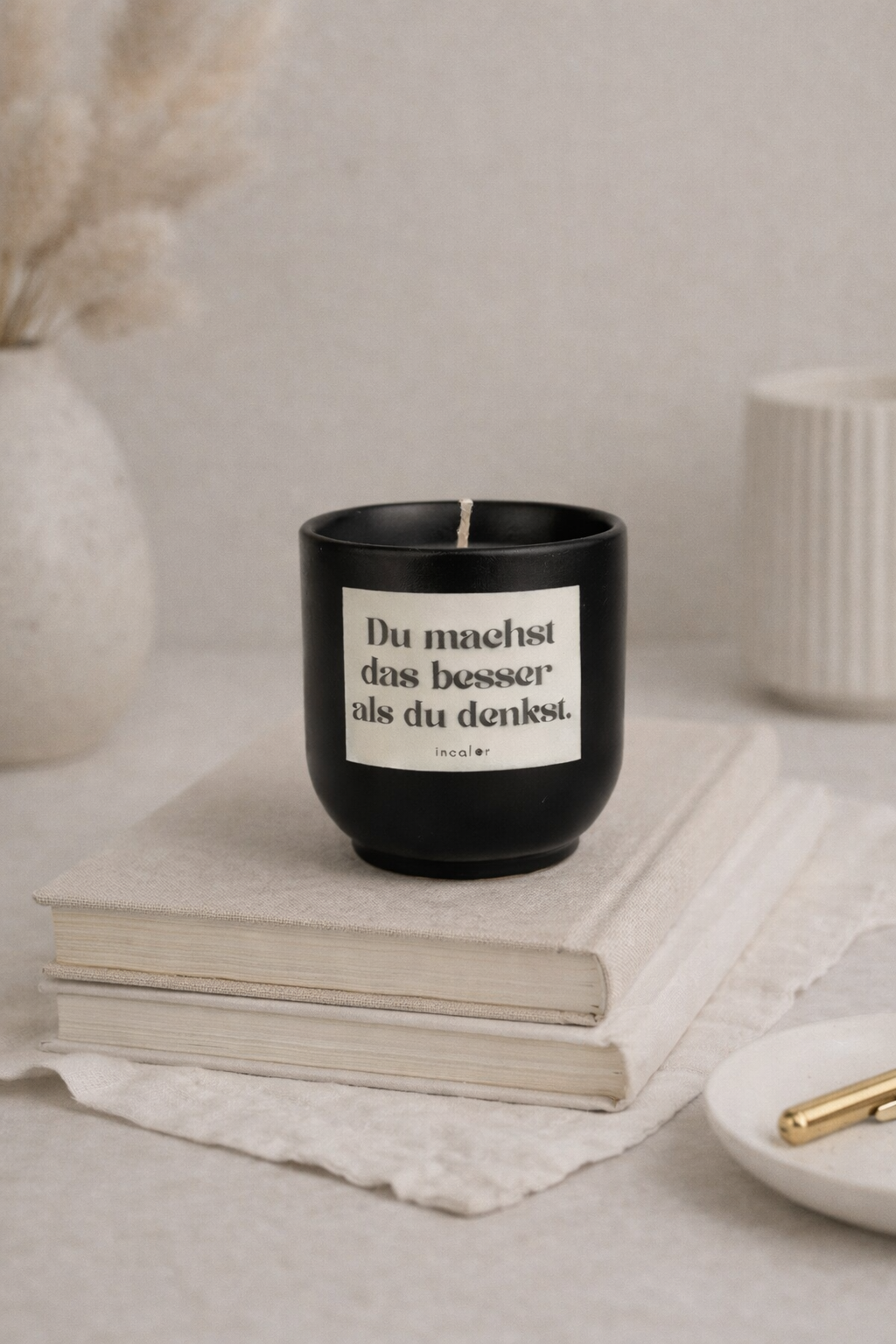 Schwarze (basalt) Kerze mit der Aufschrift ‚Du machst das besser als du denkst.‘ steht auf zwei übereinanderliegenden Büchern. Daneben befinden sich ein goldener Stift, eine kleine Schale und eine Vase mit Trockenblumen, wodurch eine ruhige, stilvolle Atmosphäre entsteht.