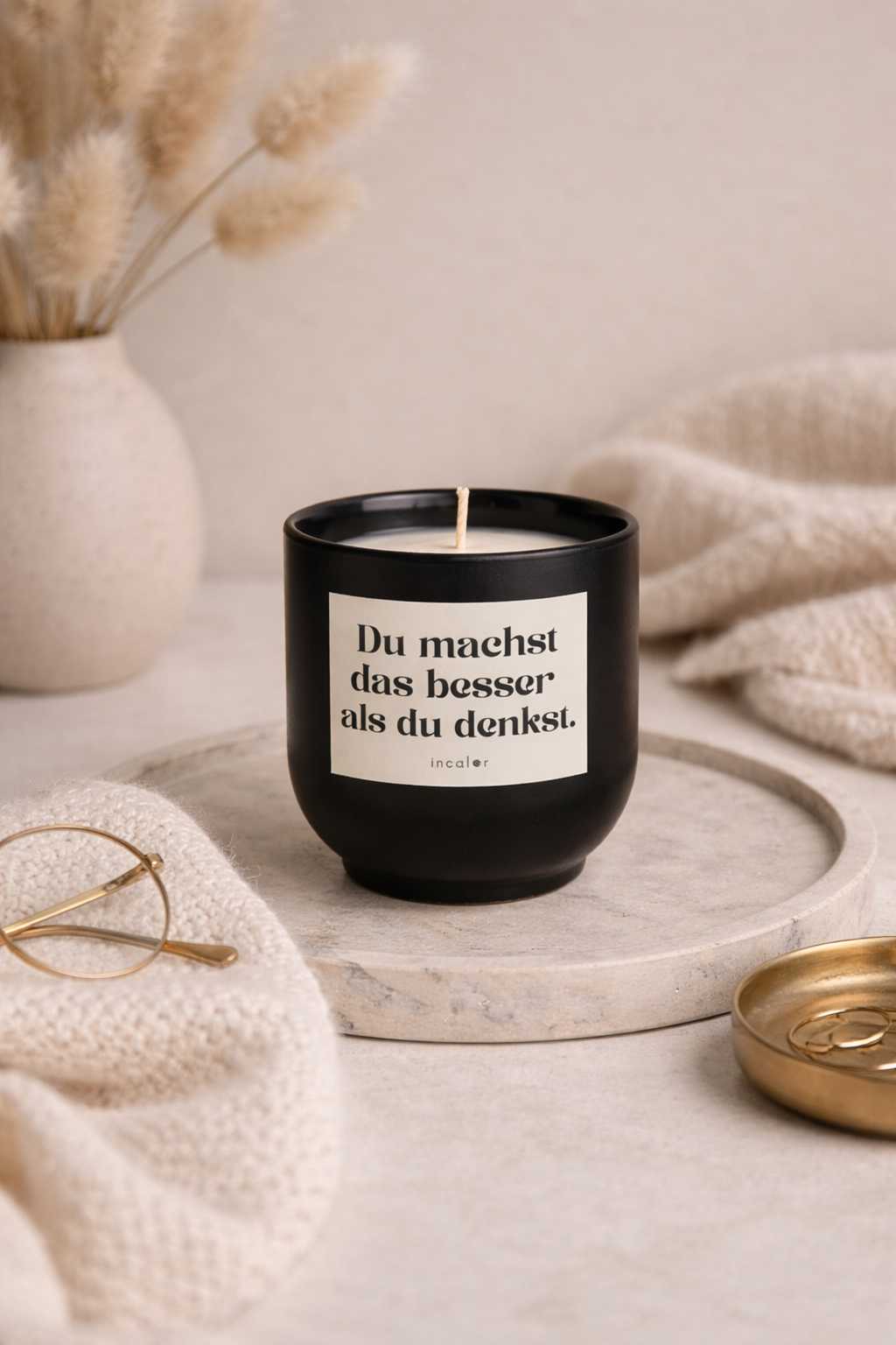 Schwarze (basalt) Duftkerze mit der Aufschrift ‚Du machst das besser als du denkst.‘ steht auf einer runden Marmorplatte. Daneben liegen eine Brille auf einem weichen Tuch und eine goldene Schale mit Ringen. Im Hintergrund sorgen eine Vase mit Trockenblumen und ein beiger Stoff für eine ruhige, warme Atmosphäre.