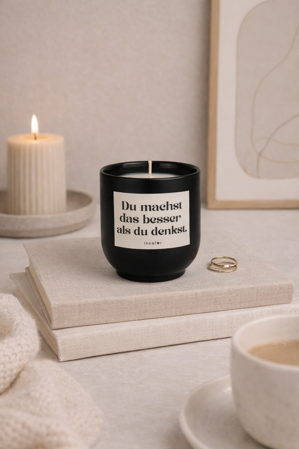 Schwarze (basalt) Kerze mit der Aufschrift ‚Du machst das besser als du denkst.‘ steht auf zwei beigen Büchern. Im Hintergrund brennt eine geriffelte Kerze, daneben liegen goldene Ringe und eine Kaffeetasse im Vordergrund – alles in einer ruhigen, warmen Wohnatmosphäre arrangiert.