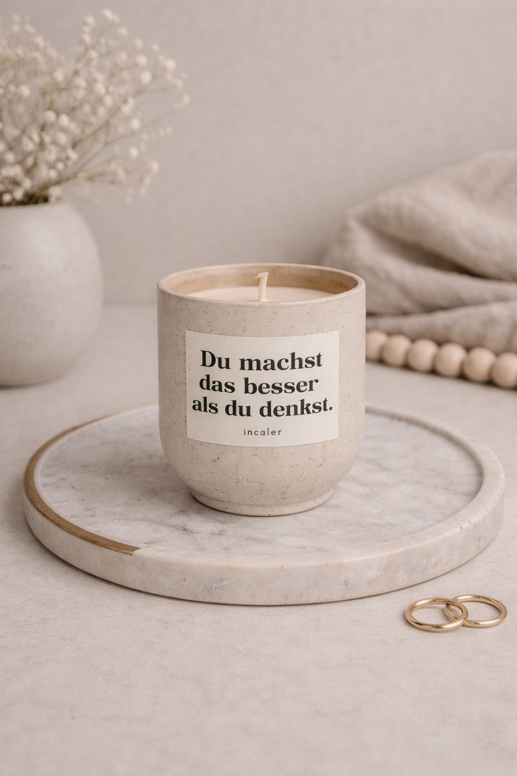 Beige (sahara) Kerze mit der Aufschrift ‚Du machst das besser als du denkst.‘ steht auf einem runden Marmortablett. Daneben liegen goldene Ringe, eine Stoffdecke und eine Vase mit zarten Trockenblumen, wodurch eine ruhige, elegante Atmosphäre entsteht.