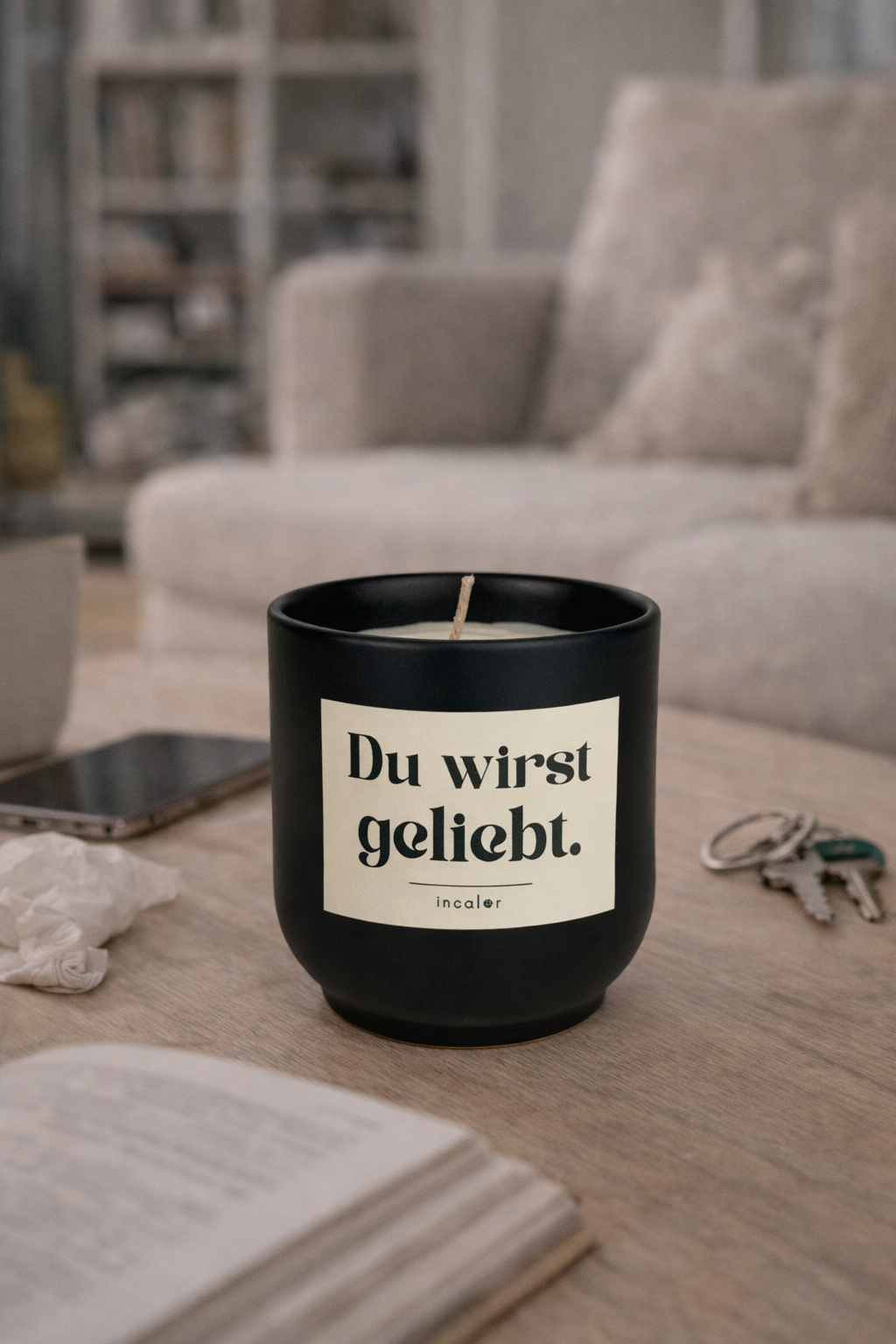 Schwarze (basalt) Kerze mit der Aufschrift ‚Du wirst geliebt.‘ steht auf einem Holztisch im Wohnzimmer. Im Hintergrund sieht man ein Sofa, daneben liegen ein Smartphone, Schlüssel und ein aufgeschlagenes Buch – die Szene wirkt gemütlich und alltagsnah.