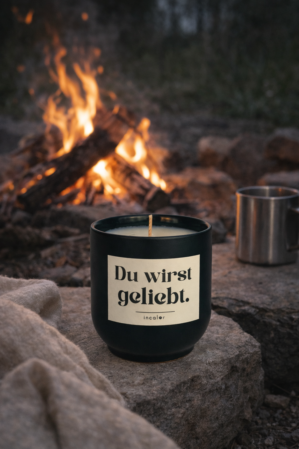 Dunkelgrüne (deep ivy) Kerze mit der Aufschrift ‚Du wirst geliebt.‘ steht auf einem Stein im Freien vor einem knisternden Lagerfeuer. Daneben befindet sich ein Metallbecher, die Szene wirkt warm, ruhig und naturnah.