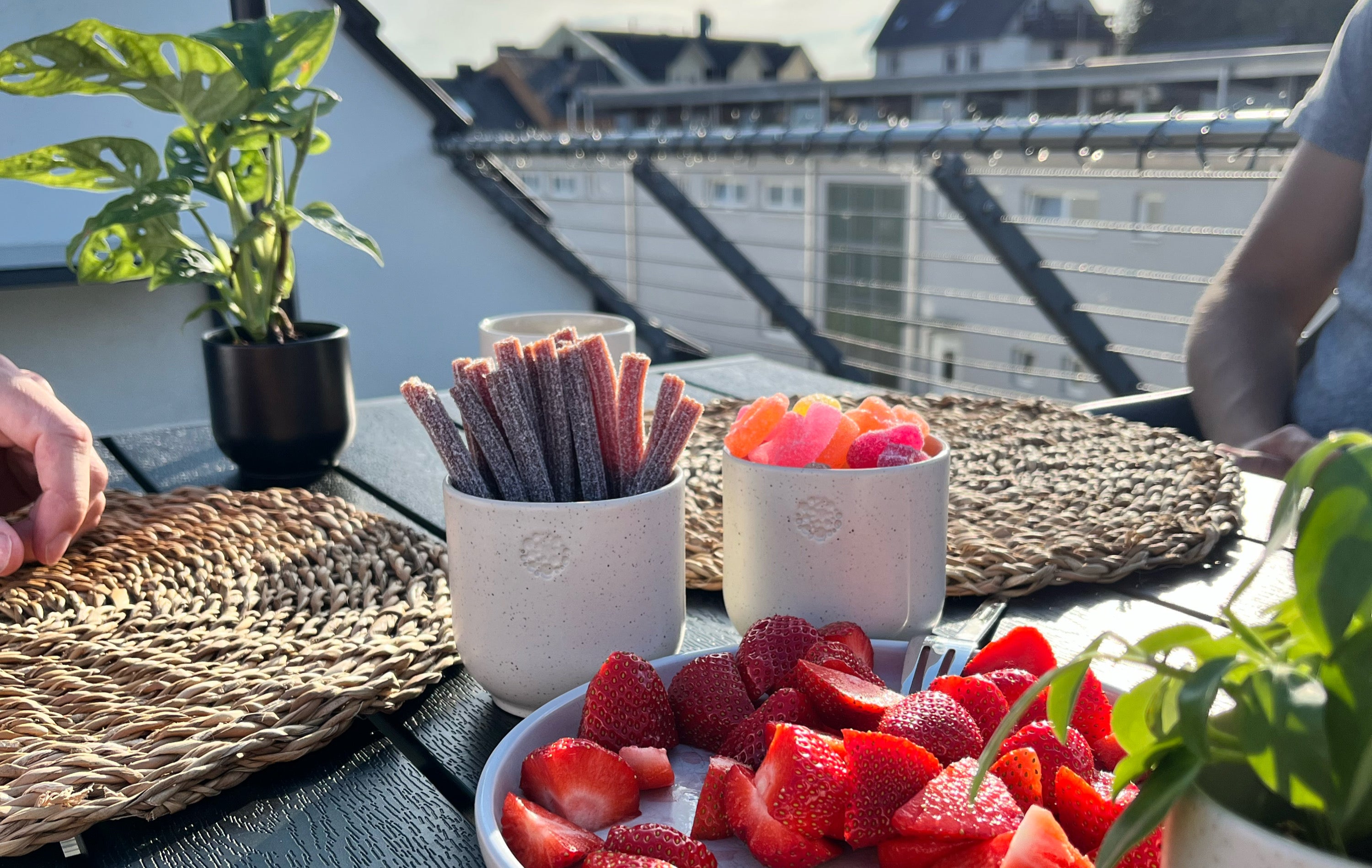 Es ist ein gedeckter Tisch zu sehen, der auf einer Terrasse steht. Auf dem Tisch steht im Hintergrund ein Becher in der Farbe basalt, der als Blumentopf genutzt wird. Im Mittelgrund sind zwei Becher in der Farbe sahara zu sehen, die jeweils mit Gummibärchen gefüllt sind. Vor den Bechern, allerdings nicht fokussiert, steht ein Teller mit aufgeschnittenen Erdbeeren. 