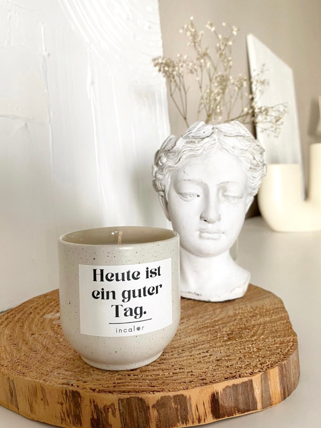 Eine cremeweiße Keramikkerze mit schwarzer Aufschrift steht auf einer Baumscheibe aus Holz. Der Text auf der Kerze lautet „Heute ist ein guter Tag.“ Daneben steht eine weiße, büstenartige Skulptur, die einen Frauenkopf darstellt. Hinter der Skulptur ist ein Zweig mit getrockneten Gänseblümchen zu sehen. Der Hintergrund ist weiß gestrichen.