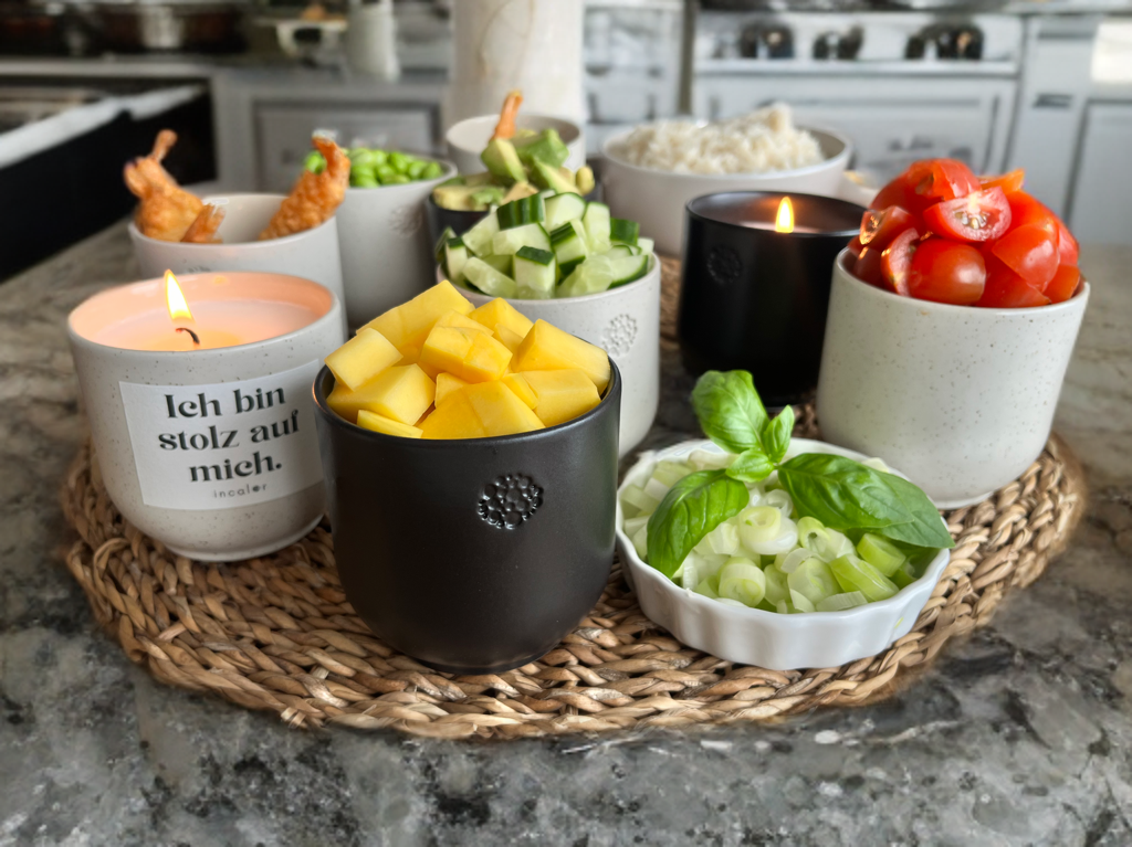 Ein Blick von oben zeigt eine Anordnung von kleinen incalor Gefäßen auf einem runden, geflochtenen Untersetzer. Jede Schale enthält eine andere Zutat, darunter gewürfelte Mango, Kirschtomaten, Edamame, Reis, Gurken und Frühlingszwiebeln mit Basilikum. Zwei Kerzen sind ebenfalls zu sehen. Eine davon ist angezündet und trägt den  Spruch „Ich bin stolz auf mich.“. Die Szene ist auf einer marmorierten Kücheninsel arrangiert.