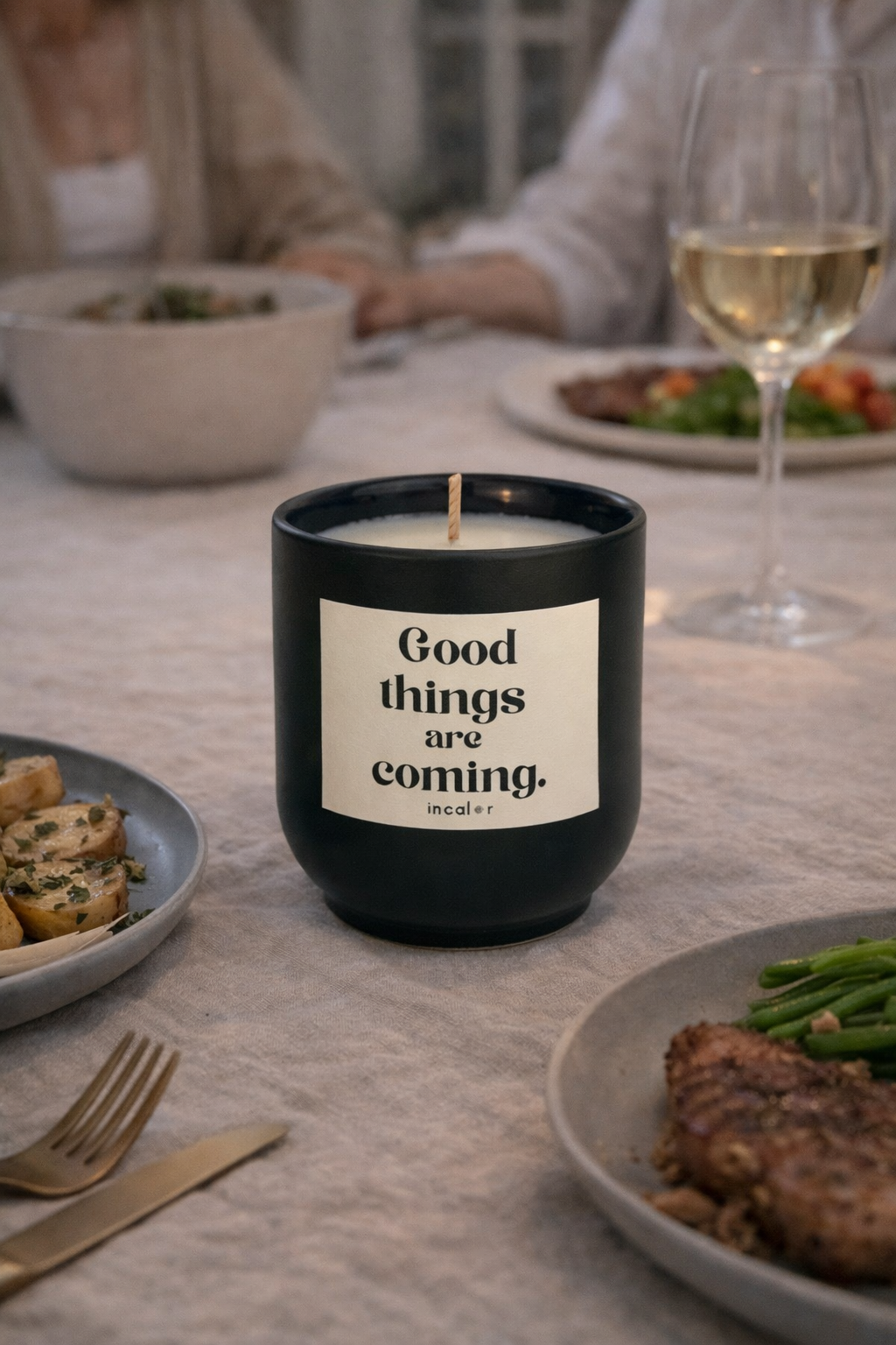 Dunkelgrüne (deep ivy) Kerze mit der Aufschrift ‚Good things are coming‘ steht auf einem gedeckten Esstisch mit Tellern, Besteck und einem Glas Weißwein. Die Szene vermittelt eine gemütliche, gesellige Atmosphäre.