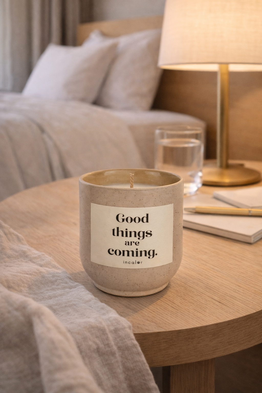 Beige (sahara), gesprenkelte Kerze mit der Aufschrift ‚Good things are coming‘ steht auf einem Holztisch neben einer Lampe, einem Glas Wasser und einem Notizbuch. Die warme Beleuchtung sorgt für eine gemütliche, entspannte Atmosphäre.
