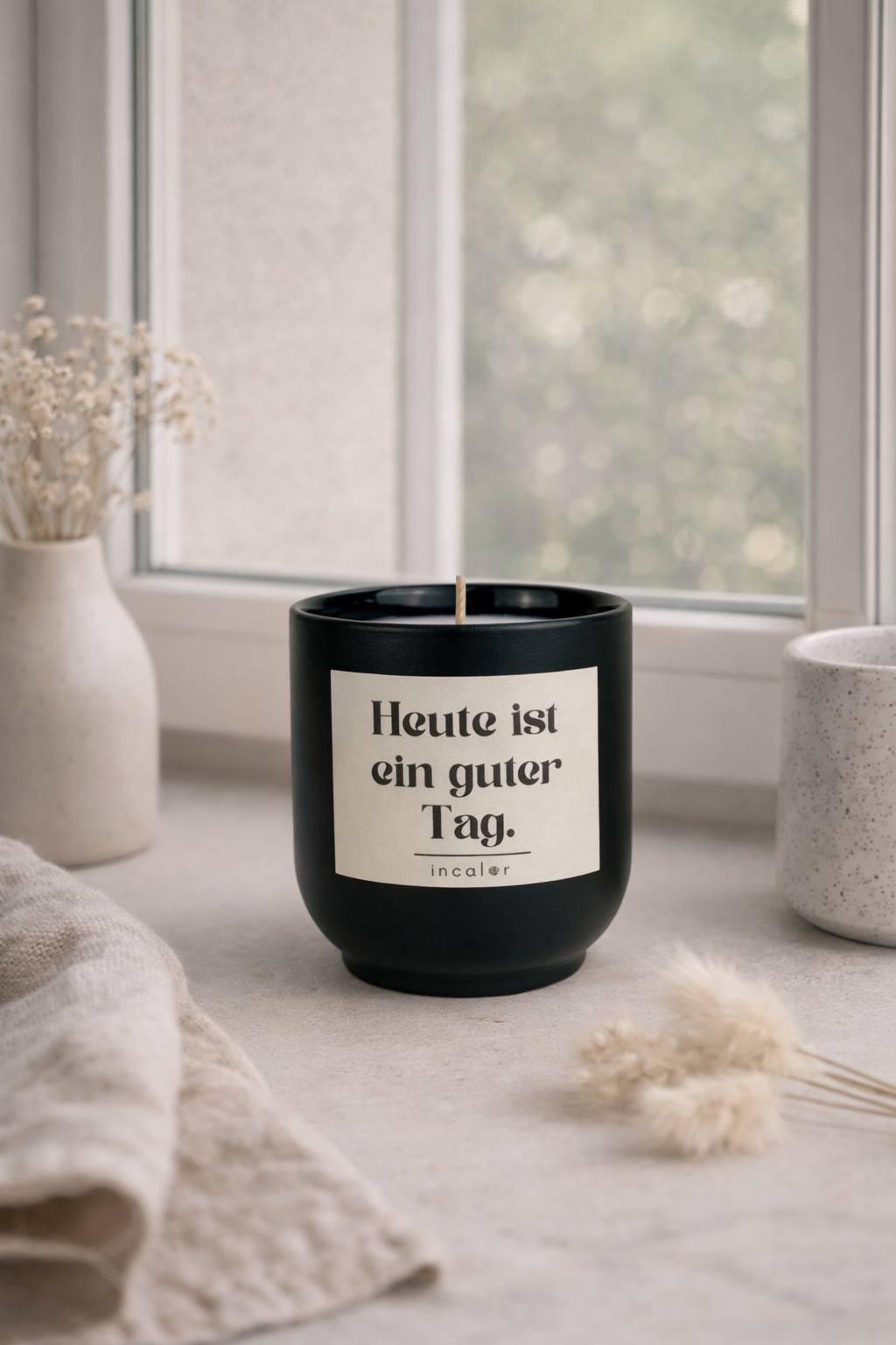 Dunkelgrüne (deep ivy) Kerzen-Tasse mit der Aufschrift ‚Heute ist ein guter Tag‘ steht auf einer hellen Fensterbank. Im Hintergrund sorgen ein Fenster, eine Vase mit Trockenblumen und weiche Textilien für eine ruhige, gemütliche Atmosphäre.