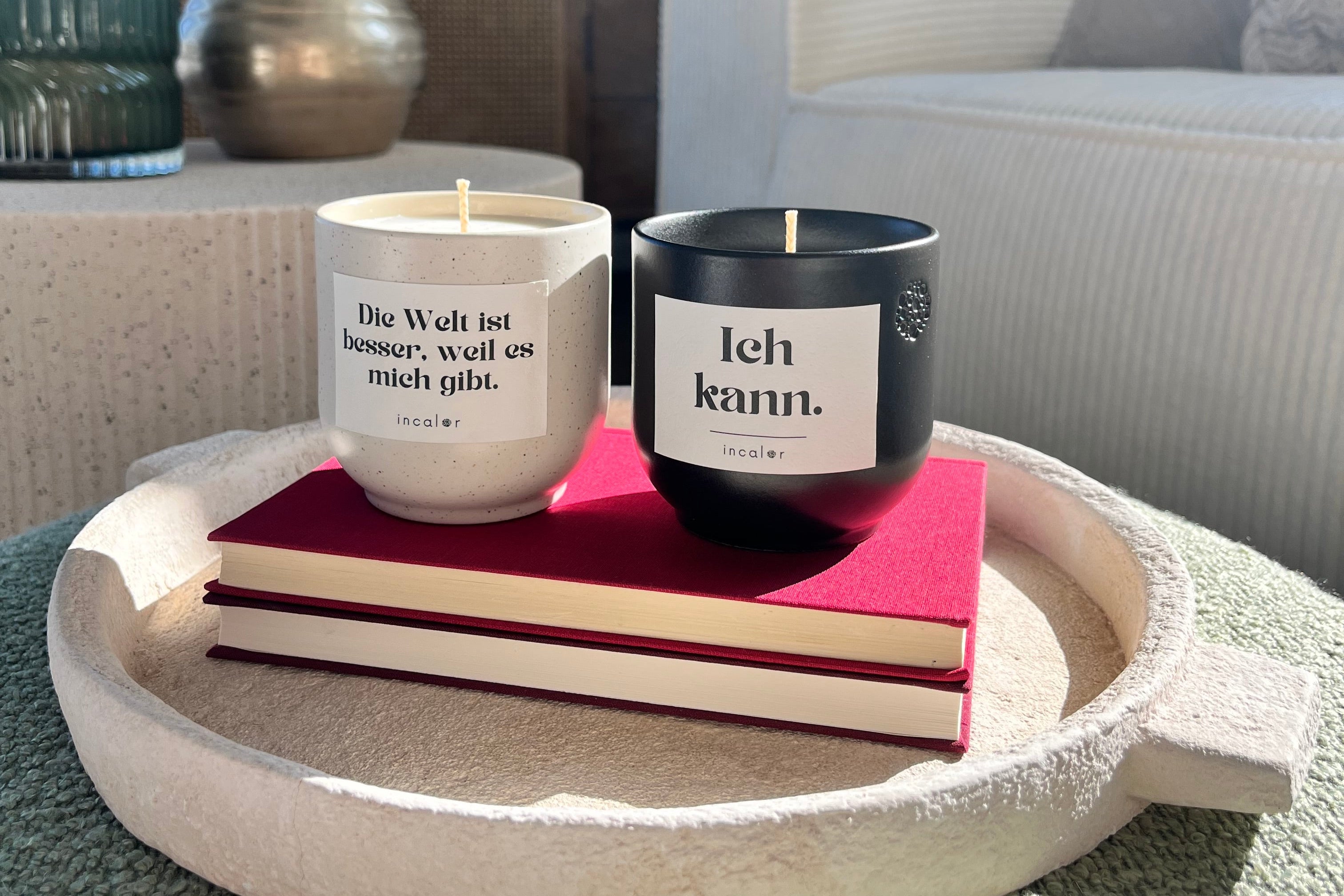 Zwei Kerzen stehen nebeneinander auf zwei roten Büchern in einem runden, hellen Dekotablett. Die linke Kerze ist weiß und trägt den Aufdruck „Die Welt ist besser, weil es mich gibt.“ Die rechte Kerze ist schwarz mit der Aufschrift „Ich kann.“ Im Hintergrund befinden sich ein helles Sofa mit Kissen, eine goldene Vase mit Zweigen und weitere Deko-Elemente. Sonnenlicht fällt auf die Szene und erzeugt warme, einladende Lichtreflexe.
