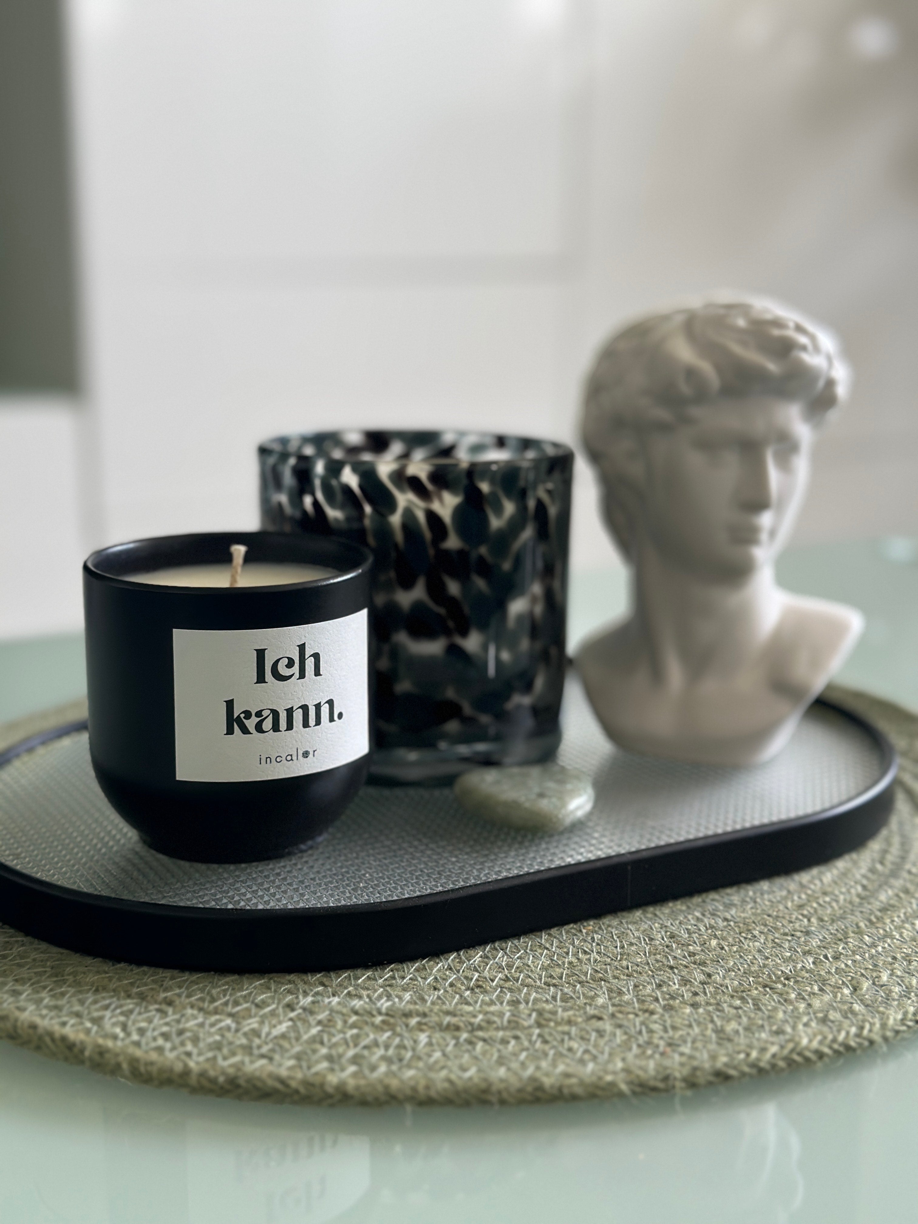 Eine Kerze in der Farbe basalt mit dem Etikett "Ich kann." steht auf einem Tablett mit weiteren Deko-Objekten: Dahinter unscharf angeordnet sind ein weißer Büstenkopf und eine Kerze in einem Glasbältnis. 