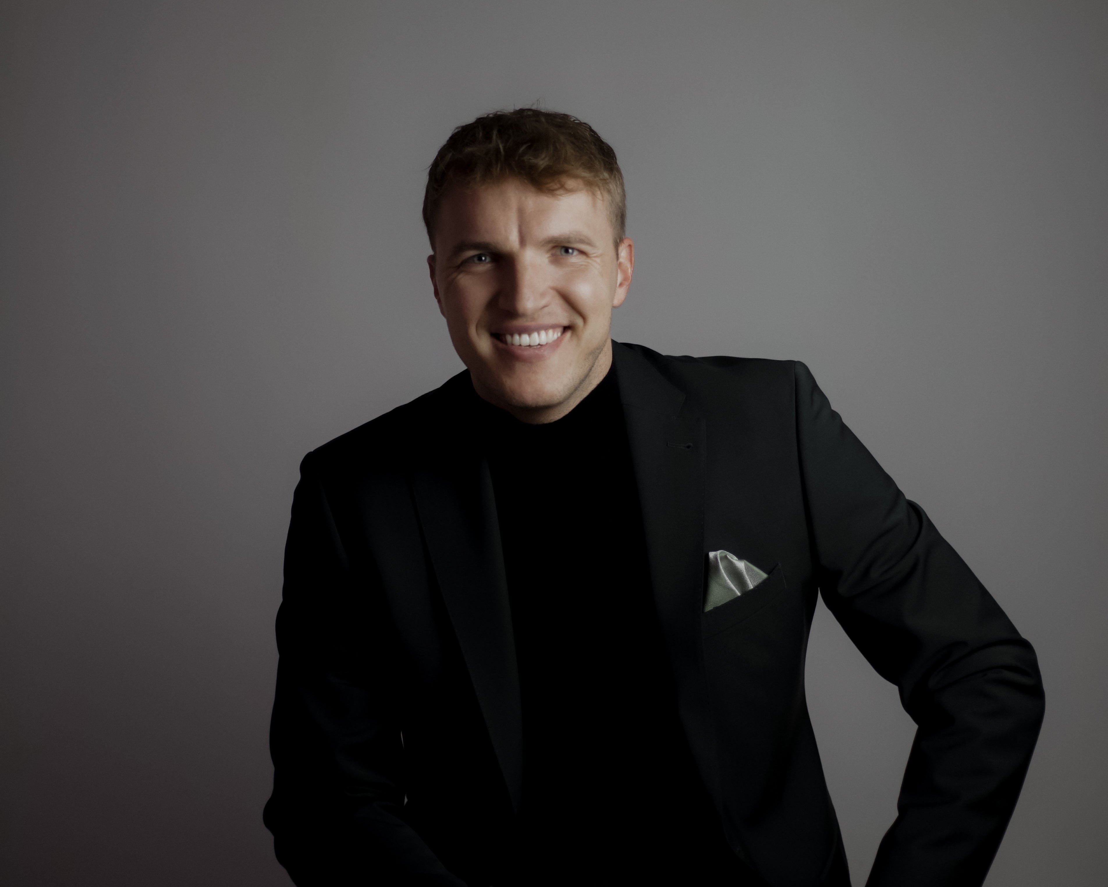 Matthias, ein junger Mann, sitzt auf einem weißen Hocker vor grauem Hintergrund. Er trägt einen eleganten schwarzen Anzug mit schwarzem Rollkragenpullover und einem hellen Einstecktuch. Er lächelt freundlich in die Kamera, seine Hände ruhen locker auf den Oberschenkeln.