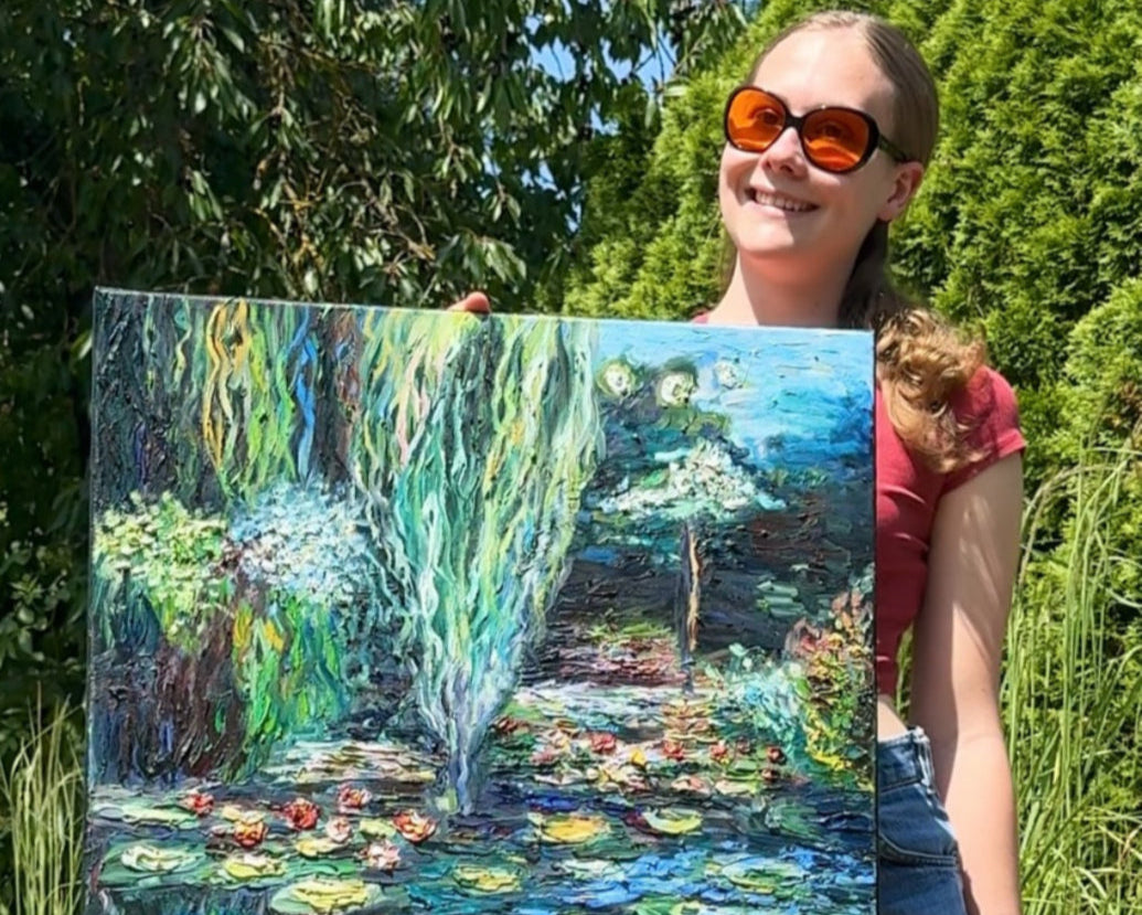 Eine junge Frau namens Jana stehe in einem Garten. Sie hat blondes Haar, das zu einem Zopf gebunden ist. Sie trägt in kurzes, rotes T-Shirt, das man kaum sieht, weil sie eine große Leinwand vor sich hält. Auf der Leinwand ist eine Wasserlandschaft mit Seerosen und wilden Pflanzen gemalt. Jana trägt eine große Brille mit organgenen Gläsern. 