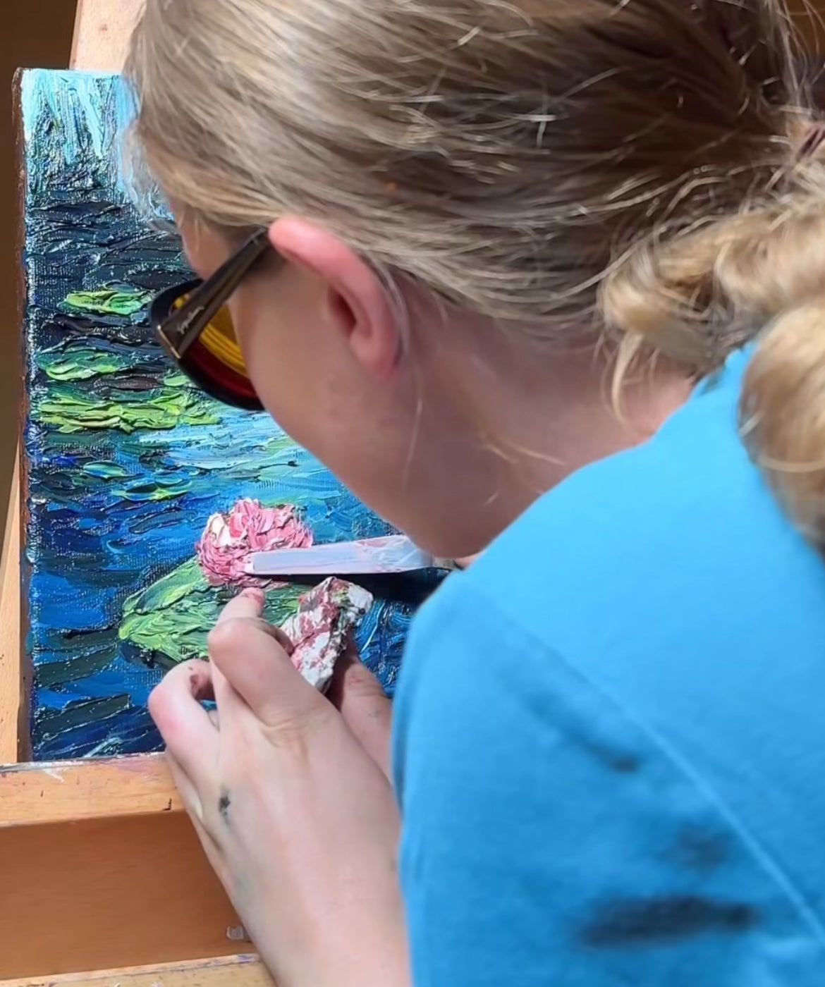 Jana sitzt mit blonden, zu einem Zopf gebundenen Haaren und Sonnenbrille  dicht vor einer Leinwand und malt mit einem Malmesser an einem Bild. Das Gemälde zeigt in kräftigen Farben einen blauen Teich mit grünen Seerosenblättern und einer rosa Seerosenblüte im Vordergrund.