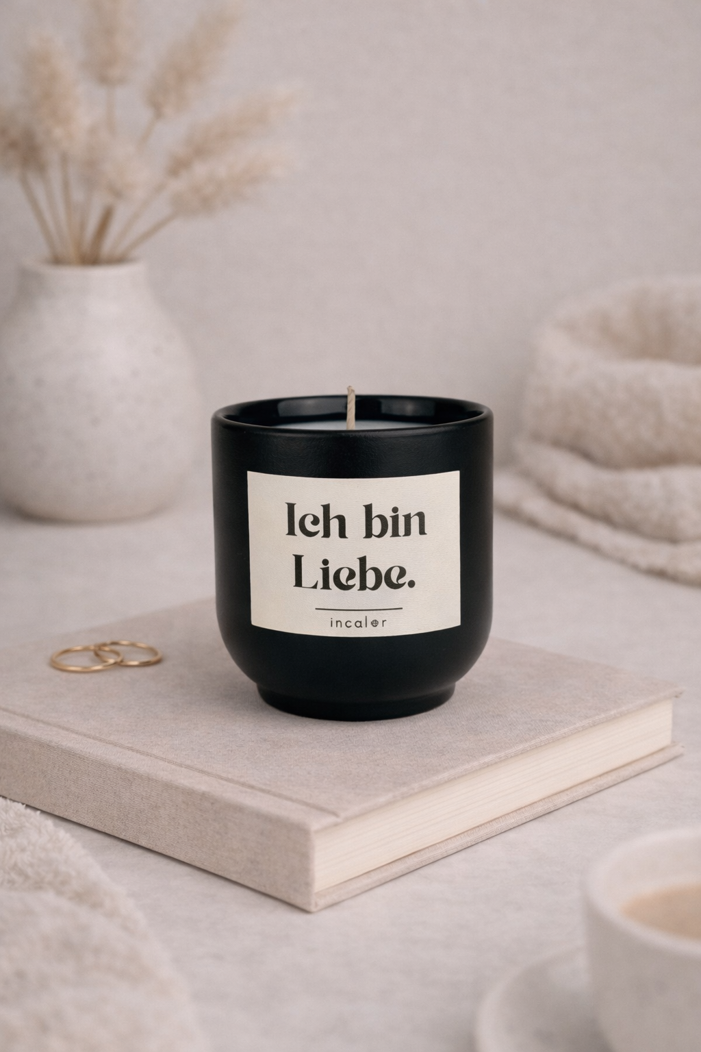 Schwarze (Basalt) Kerzen-Tasse mit der Aufschrift ‚Ich bin Liebe‘ steht auf einem beigen Buch. Daneben liegen goldene Ringe, im Hintergrund sorgen eine Vase mit Trockenblumen und weiche Textilien für eine ruhige, warme Atmosphäre.