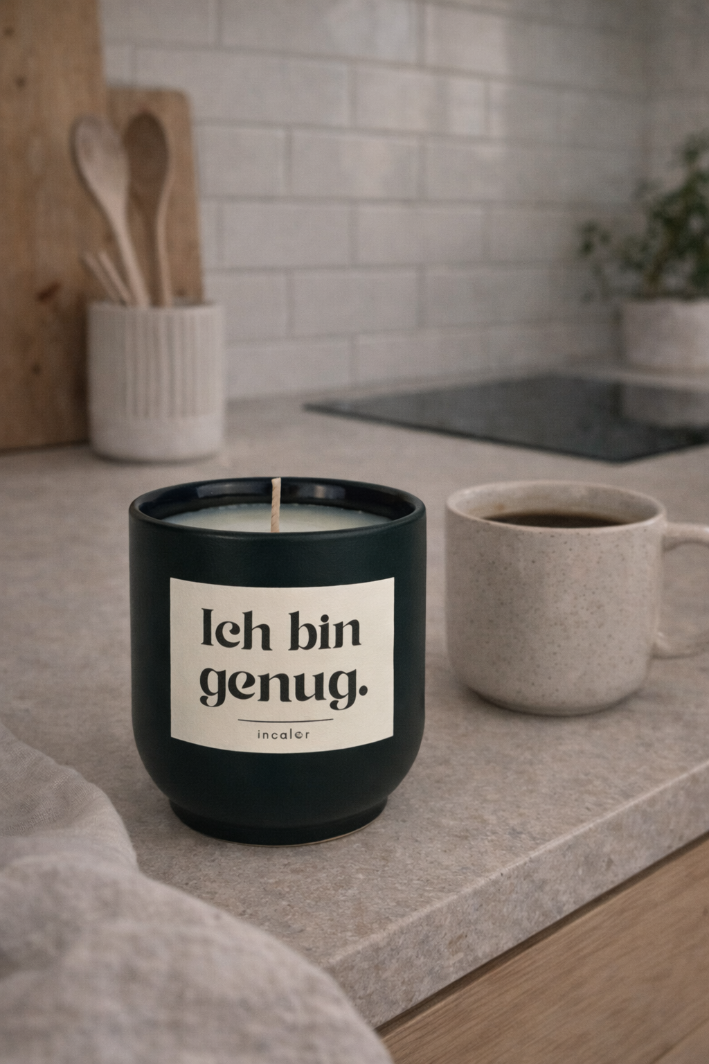 Dunkelgrüne (deep ivy) Kerze mit der Aufschrift ‚Ich bin genug.‘ steht auf einer Küchenarbeitsplatte. Daneben befindet sich eine Tasse Kaffee, im Hintergrund sind eine moderne Küche mit Holzutensilien und ein Kochfeld zu sehen – die Szene wirkt ruhig und gemütlich.
