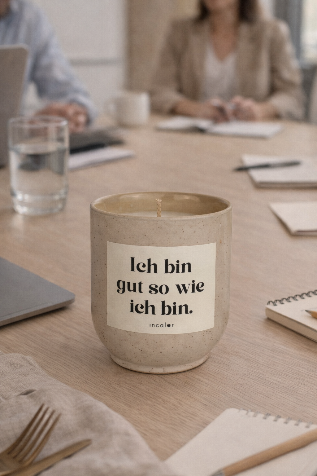 Beige (sahara) Kerze mit der Aufschrift ‚Ich bin gut so wie ich bin.‘ steht auf einem Schreibtisch. Im Hintergrund sitzen zwei Personen in einem Meeting mit Laptop, Notizbüchern und einem Glas Wasser – die Szene wirkt ruhig und modern.