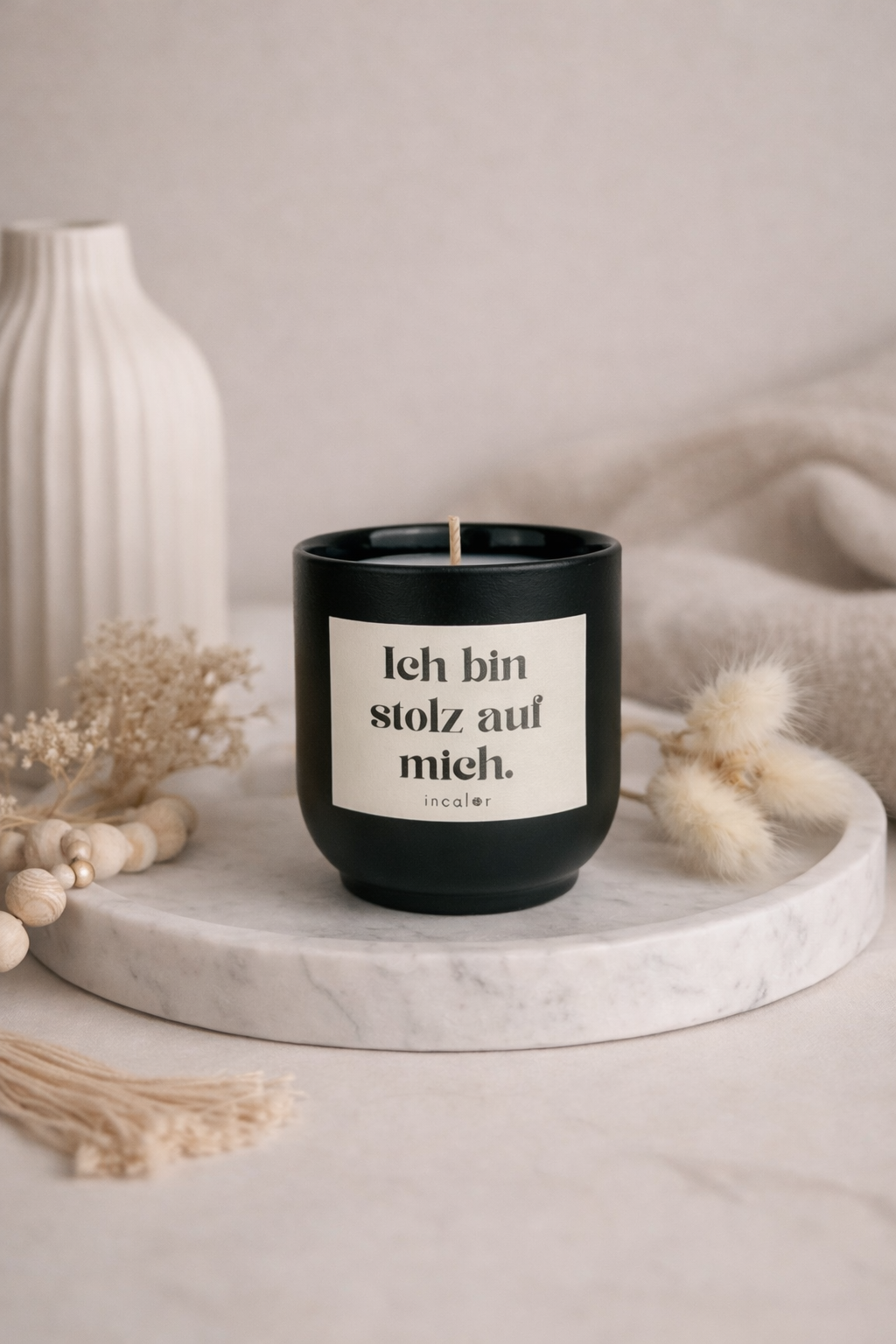 Schwarze (basalt) Duftkerze mit der Aufschrift ‚Ich bin stolz auf mich‘ steht mittig auf einem runden Marmortablett. Umgeben von einer schlichten Vase, Trockenblumen und weichen Textilien entsteht eine ruhige, minimalistische und warme Wohnatmosphäre.
