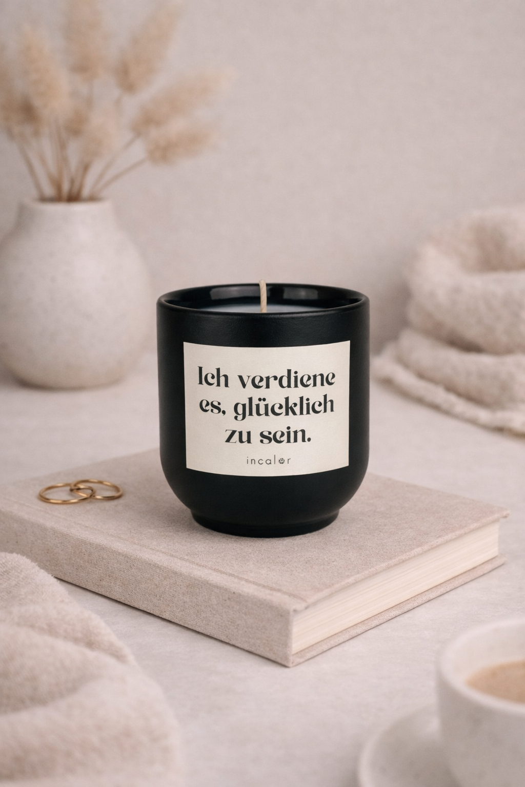 Dunkelgrüne (deep ivy) Kerzen-Tasse mit der Aufschrift ‚Ich verdiene es, glücklich zu sein‘ steht auf einem beigen Buch. Daneben liegen goldene Ringe, im Hintergrund sorgen eine Vase mit Trockenblumen und weiche Textilien für eine ruhige, warme Wohlfühlatmosphäre.