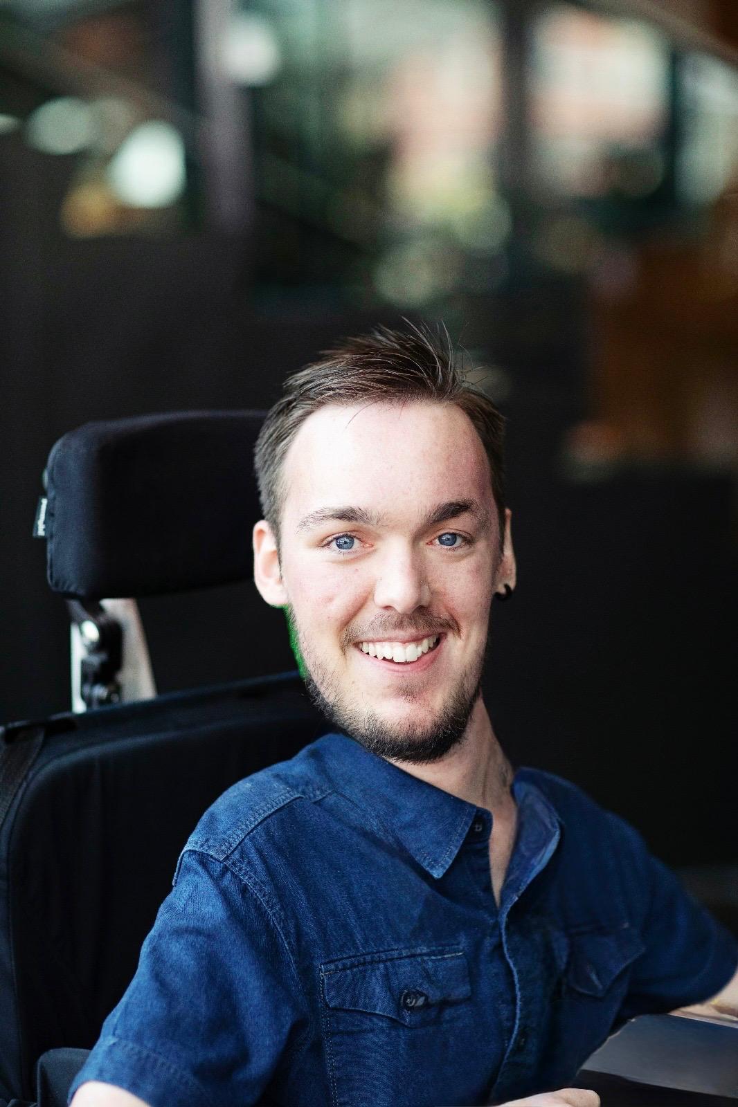 Joshua, ein junger Mann mit braunen Haaren, sitzt im Rollstuhl und lächelt freundlich in die Kamera.