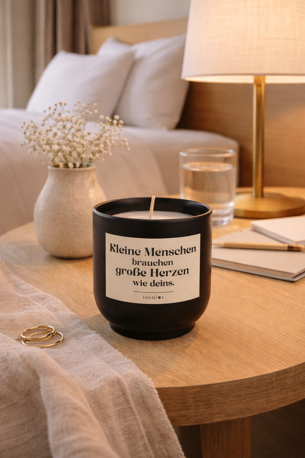 Schwarze (basalt) Kerze mit der Aufschrift ‚Kleine Menschen brauchen große Herzen wie deins‘ steht auf einem Holztisch neben einer Lampe, einem Glas Wasser, einem Notizbuch und einer Vase mit Blumen. Warmes Licht sorgt für eine gemütliche, liebevolle Atmosphäre.