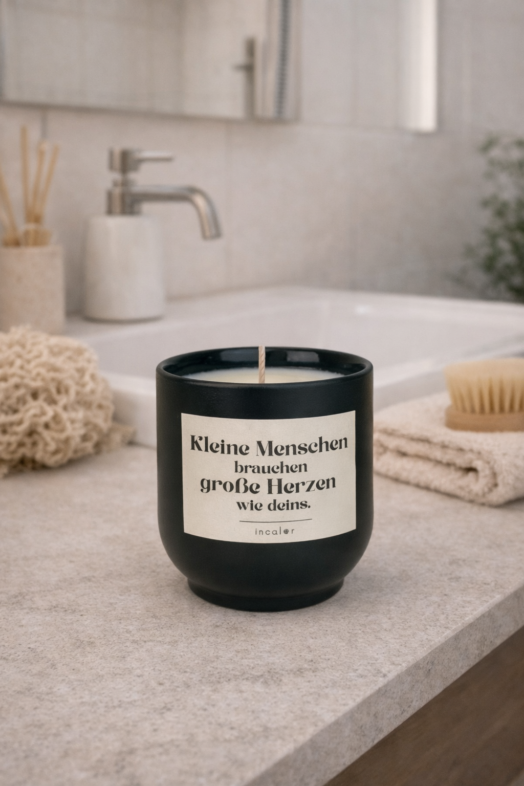 Dunklerüne (deep ivy) Kerze mit der Aufschrift ‚Kleine Menschen brauchen große Herzen wie deins‘ steht auf einem Badezimmerwaschbecken neben Seifenspender, Bürste und Handtuch. Die Szene wirkt hell, sauber und beruhigend.