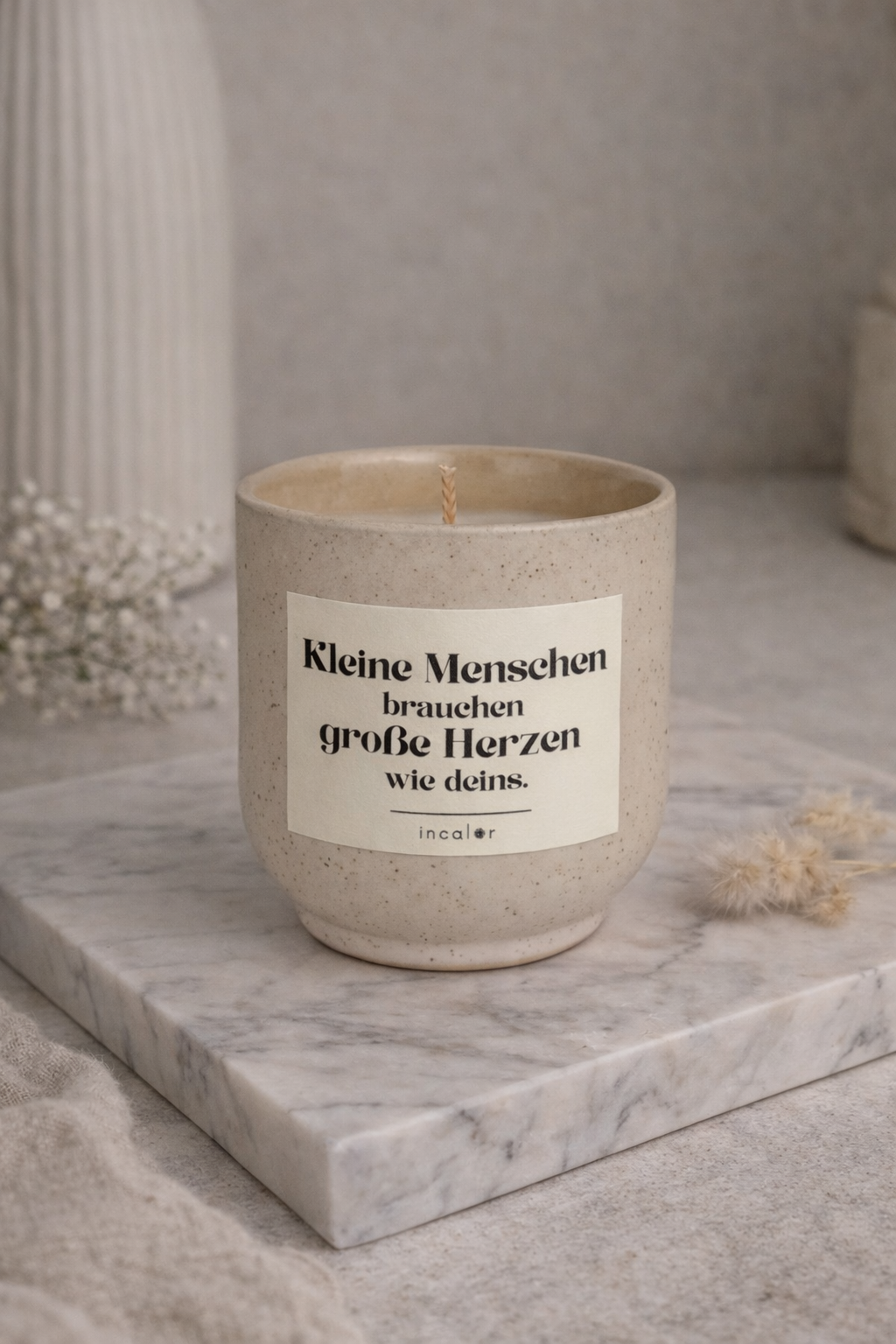 Beige (sahara), gesprenkelte Kerze mit der Aufschrift ‚Kleine Menschen brauchen große Herzen wie deins‘ steht auf einer Marmorplatte, umgeben von dezenten Deko-Elementen. Die Szene wirkt ruhig, hell und minimalistisch.