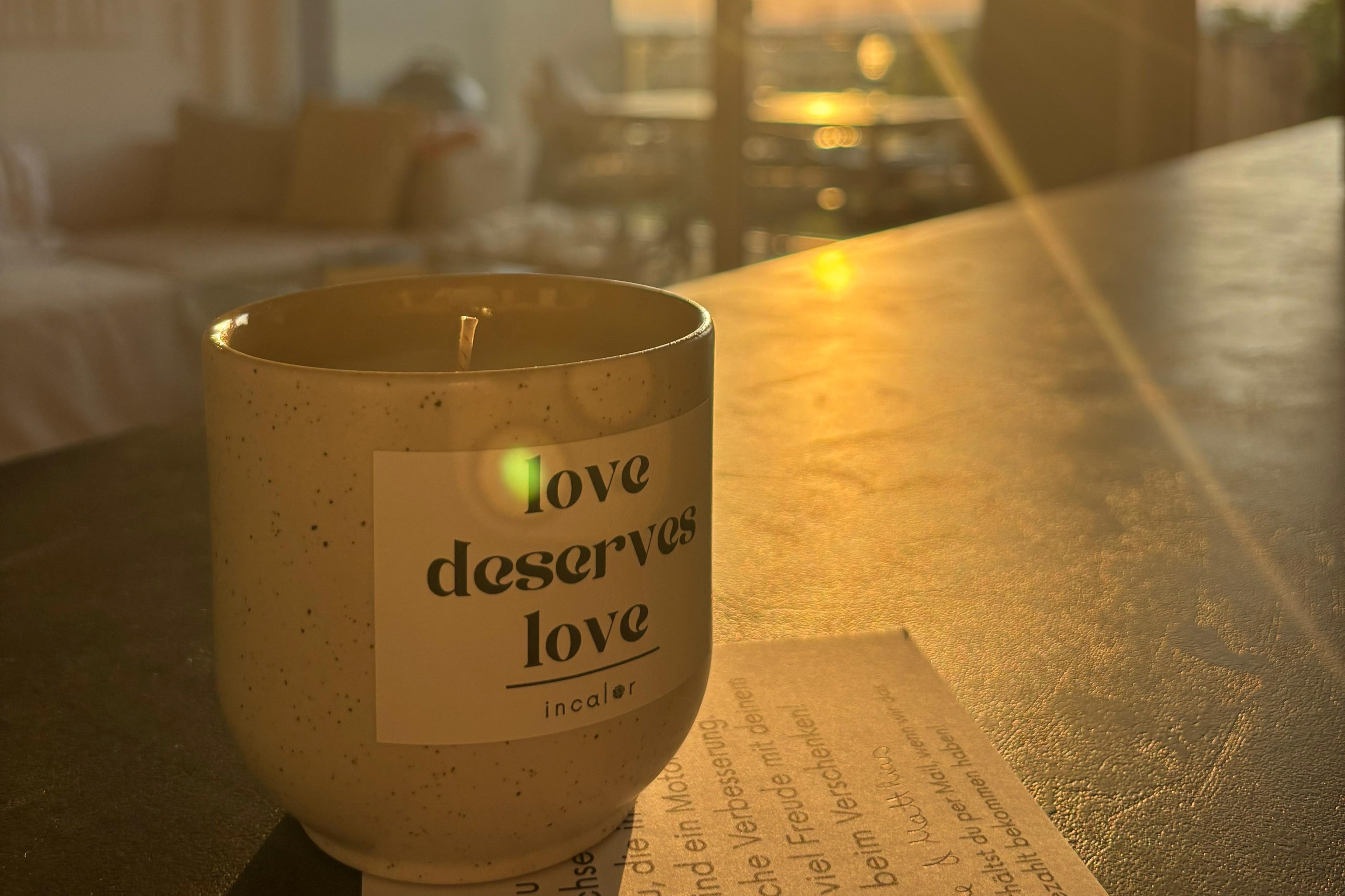 Eine beige Kerze mit dunklen Punkten aus Rost steht auf einer Schieferplatte in einem Wohnraum. Im Hintergrund scheint die Abendsonne durch ein Fenster und verursacht Lichtreflexe im Bild. Die Kerze trägt das Etikett "love deserves love", welches viereckig ist und schwarze Farbe auf weißem Hintergrund zeigt. 