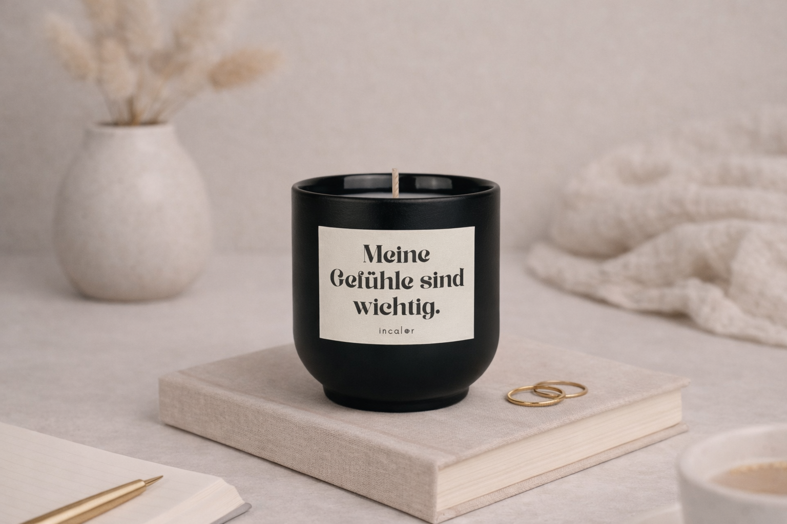 Schwarze (basalt) Duftkerze mit der Aufschrift ‚Meine Gefühle sind wichtig‘ steht auf einem geschlossenen Buch. Daneben liegen ein Notizbuch mit Stift, zwei goldene Ringe sowie eine Vase mit Trockenblumen – in einer ruhigen, warmen und minimalistischen Wohnatmosphäre.