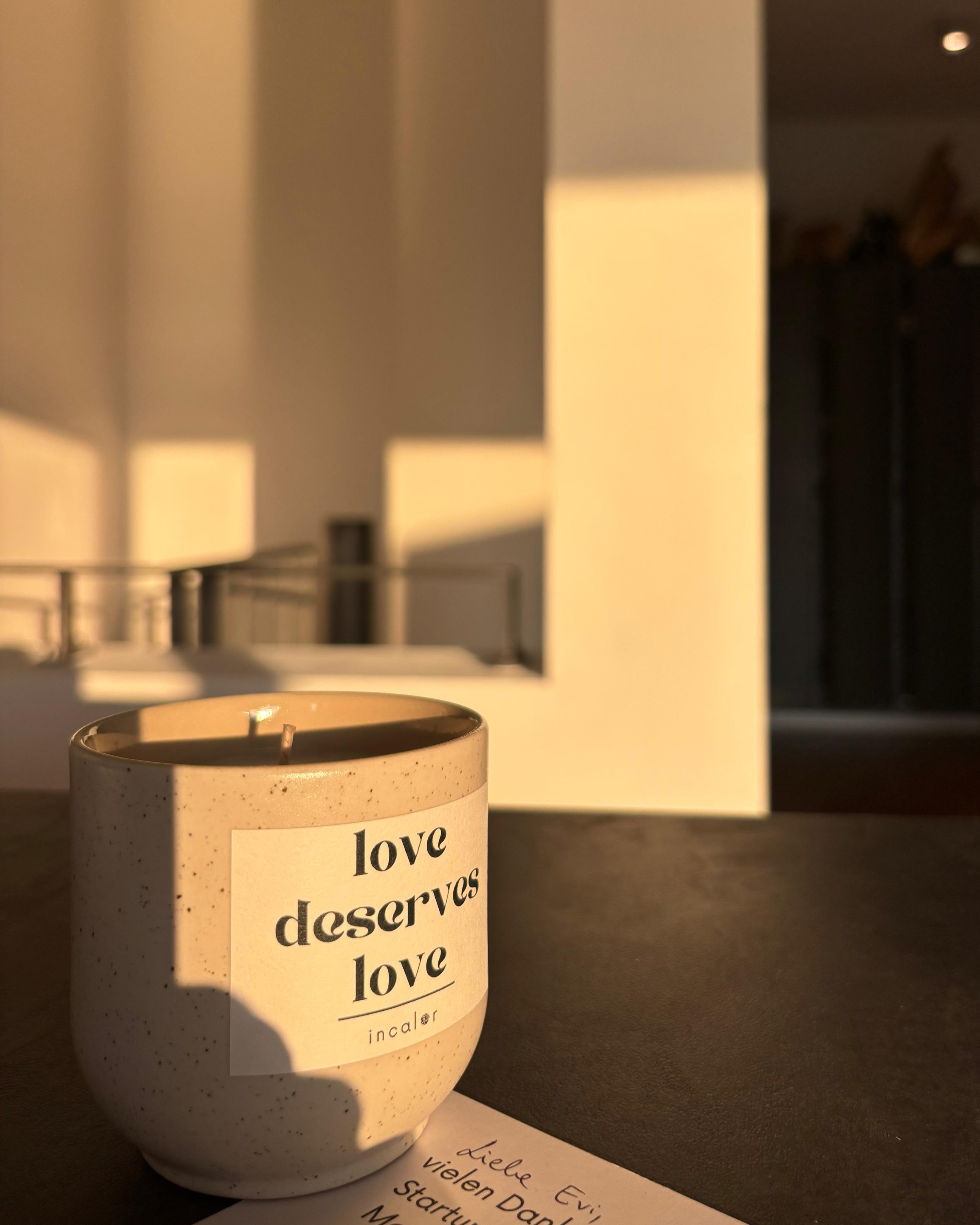 Eine cremefarbene Kerze mit dunklen Sprenkeln steht auf einem dunklen Tisch. Auf der Kerze klebt ein weißes Etikett mit dem schwarzen Text „love deserves love“ und darunter der Markenname „incalor“. Die Kerze wird von der untergehenden Sonne angestrahlt, die einen langen, warmen Lichtstreifen auf eine helle Wand im Hintergrund wirft. Im Vordergrund liegt ein Teil eines beschriebenen Zettels.