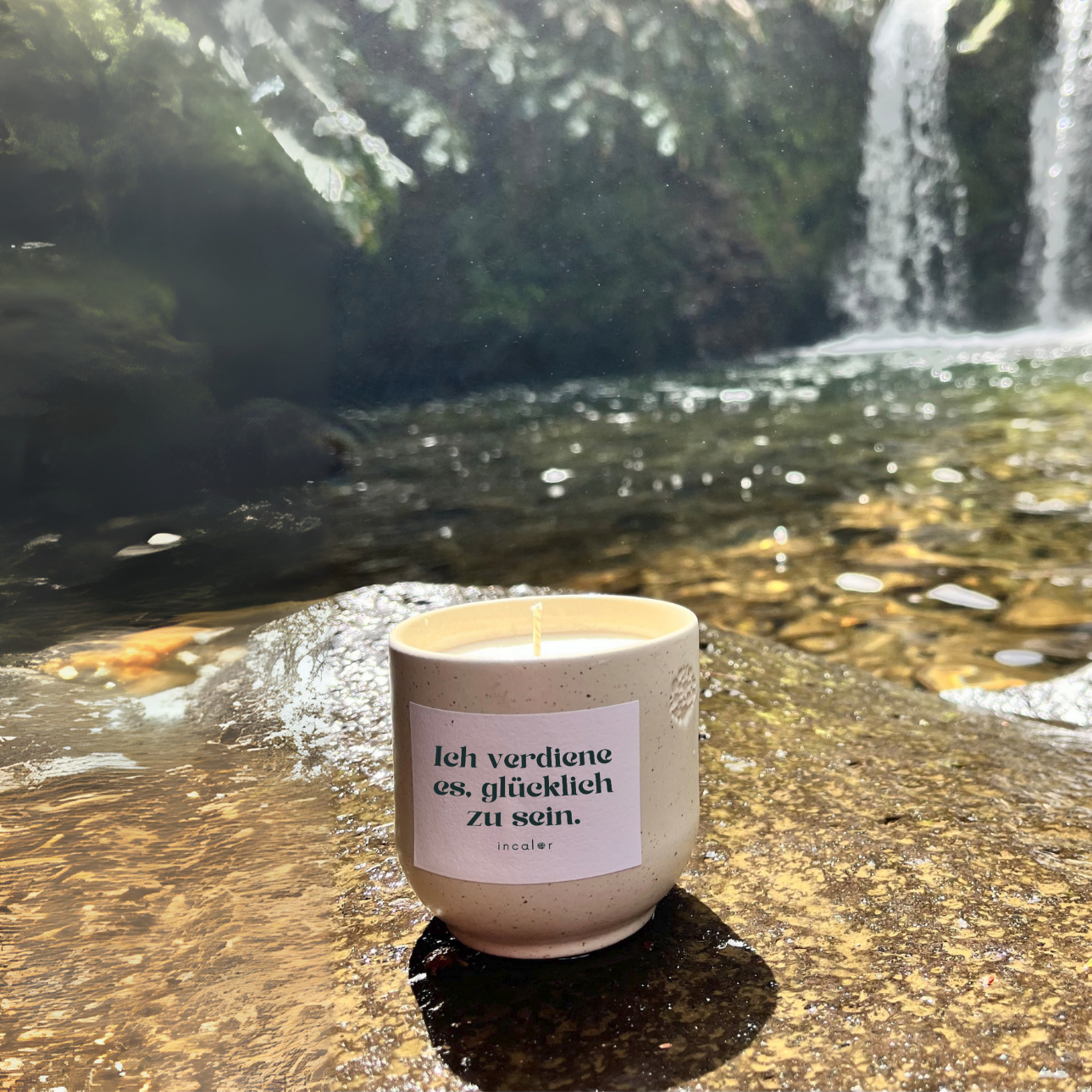 Kerze aus beige Keramik (Farbton sahara) mit Label der Affirmation "Ich verdiene es, glücklich zu sein.", stilvoll platziert auf Naturstein mit einem Wasserfall im Hintergrund – hochwertige Designkerze für modernes, minimalistisches Wohndesign.