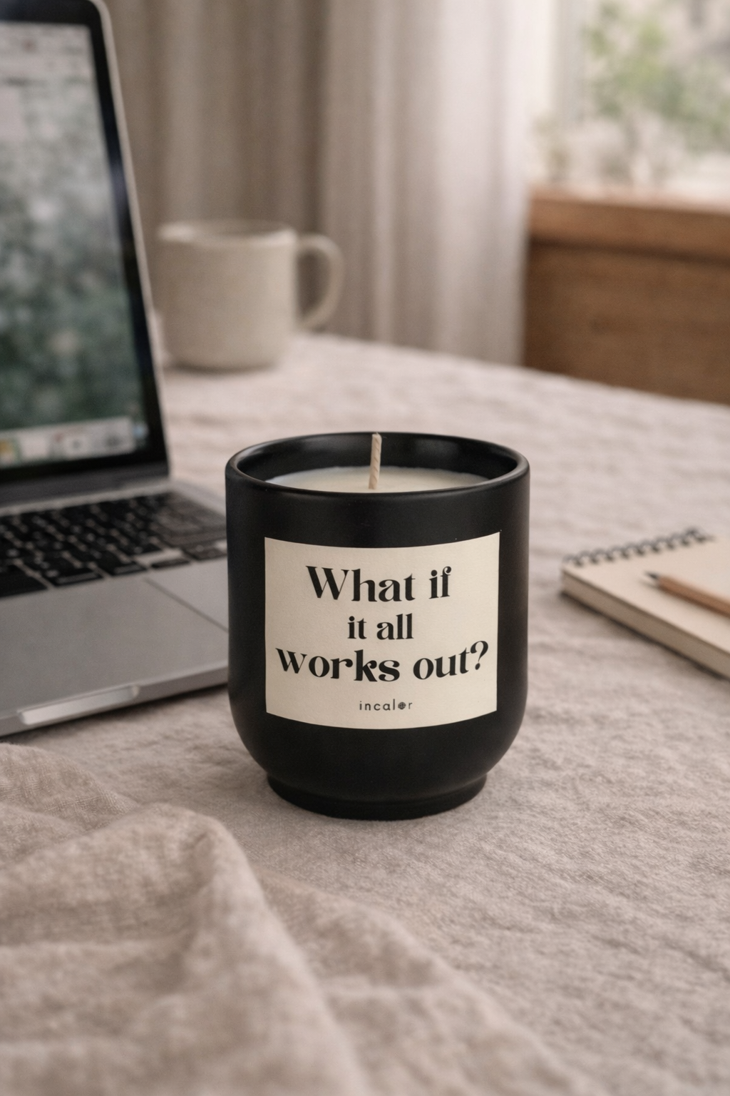 Schwarze (basalt) Kerze mit der Aufschrift ‚What if it all works out?‘ steht auf einem Tisch neben einem Laptop, einer Tasse und einem Notizbuch. Die Szene vermittelt eine ruhige, produktive und inspirierende Arbeitsatmosphäre.