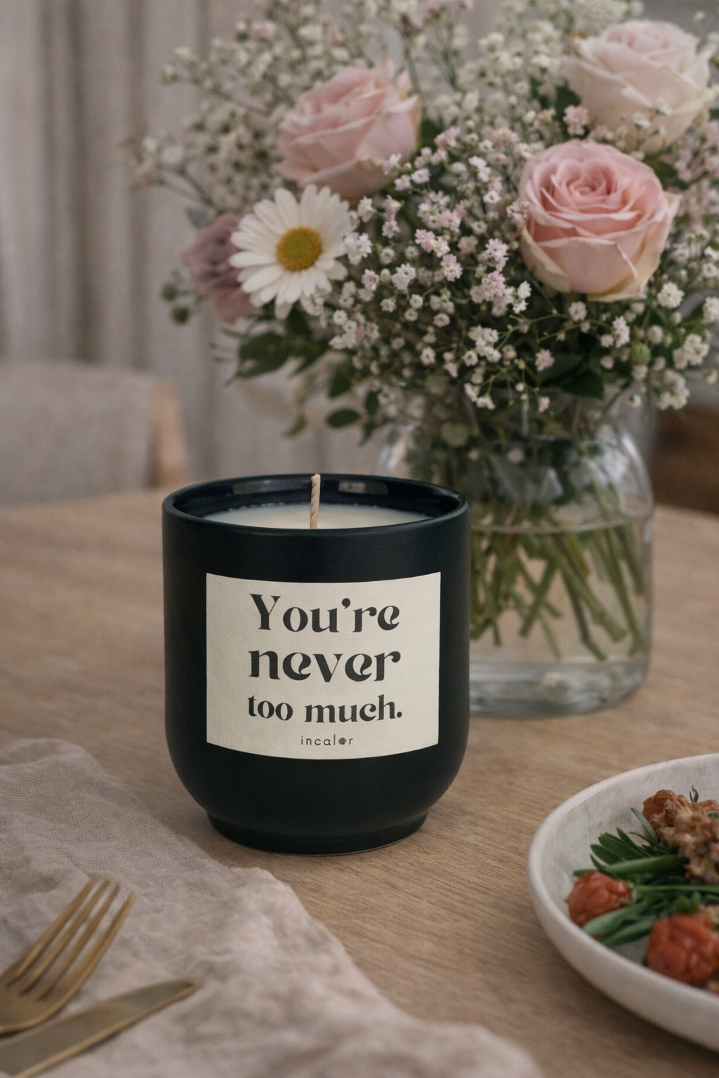 Dunkelgrüne (deep ivy) Kerze mit der Aufschrift ‚You’re never too much.‘ steht auf einem gedeckten Tisch. Im Hintergrund befindet sich ein Blumenstrauß mit Rosen und kleinen Blüten in einer Glasvase, daneben Teller und Besteck, was eine warme, einladende Atmosphäre schafft.