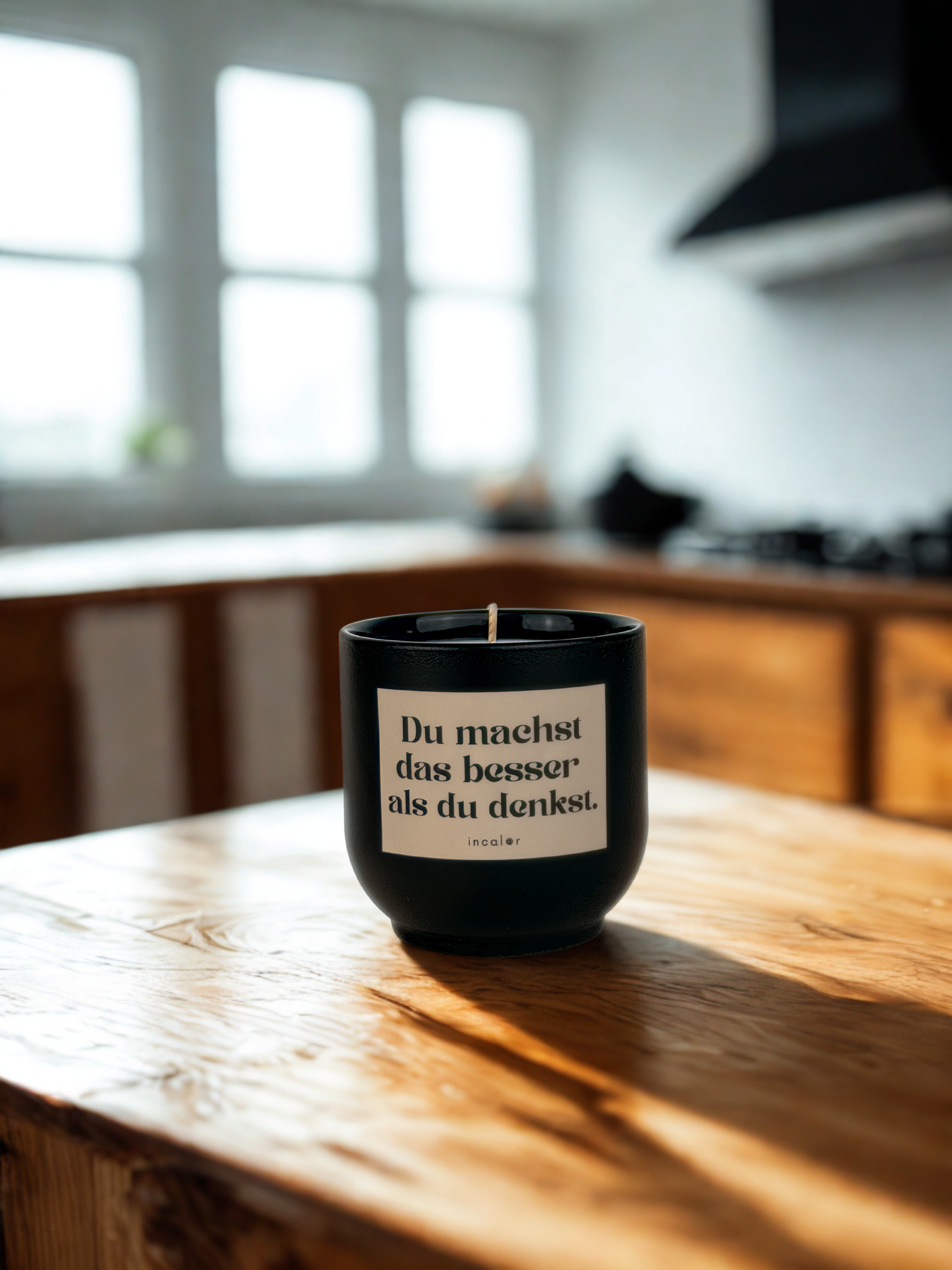 Dunkelgrüne (deep ivy) Duftkerze mit der Aufschrift „Du machst das besser als du denkst.“ steht auf einem hellen Holztisch in einer modernen Küche mit großen Fenstern im Hintergrund.