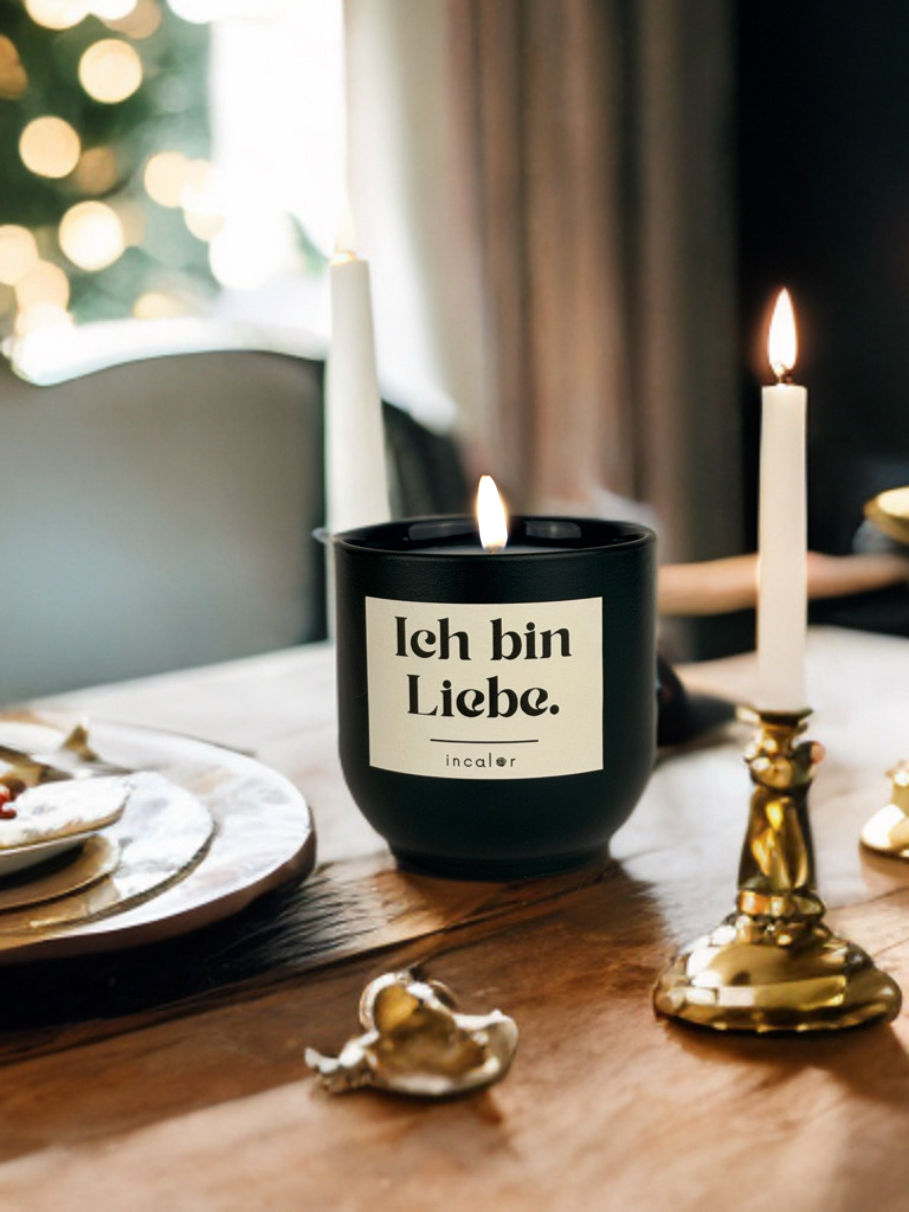 Dunkelgrüne (deep ivy) Duftkerze mit brennender Flamme steht auf einem gedeckten Holztisch. Auf dem Etikett steht „Ich bin Liebe.“ mit dem Markennamen „incalor“. Um sie herum befinden sich goldene Kerzenhalter mit weißen Stabkerzen, Geschirr und warme Lichtreflexe. Im Hintergrund sorgen unscharfe Lichter eines Weihnachtsbaums für eine festliche, gemütliche Stimmung.
