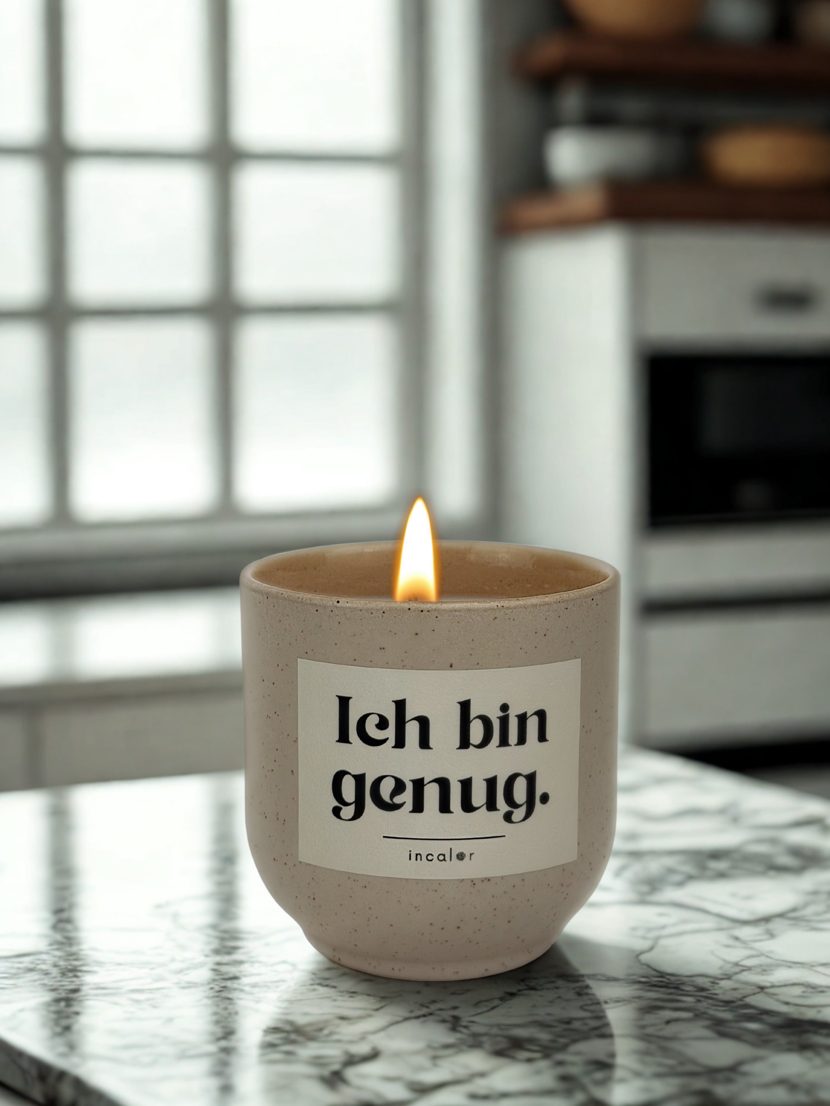 Beige (sahara) Duftkerze mit brennender Flamme steht auf einer marmorierten Arbeitsplatte in einer hellen Küche. Auf dem Etikett steht „Ich bin genug.“ mit dem Markennamen „incalor“. Im Hintergrund sind große Fenster und Küchenschränke unscharf zu erkennen, was eine ruhige, minimalistische Atmosphäre vermittelt.