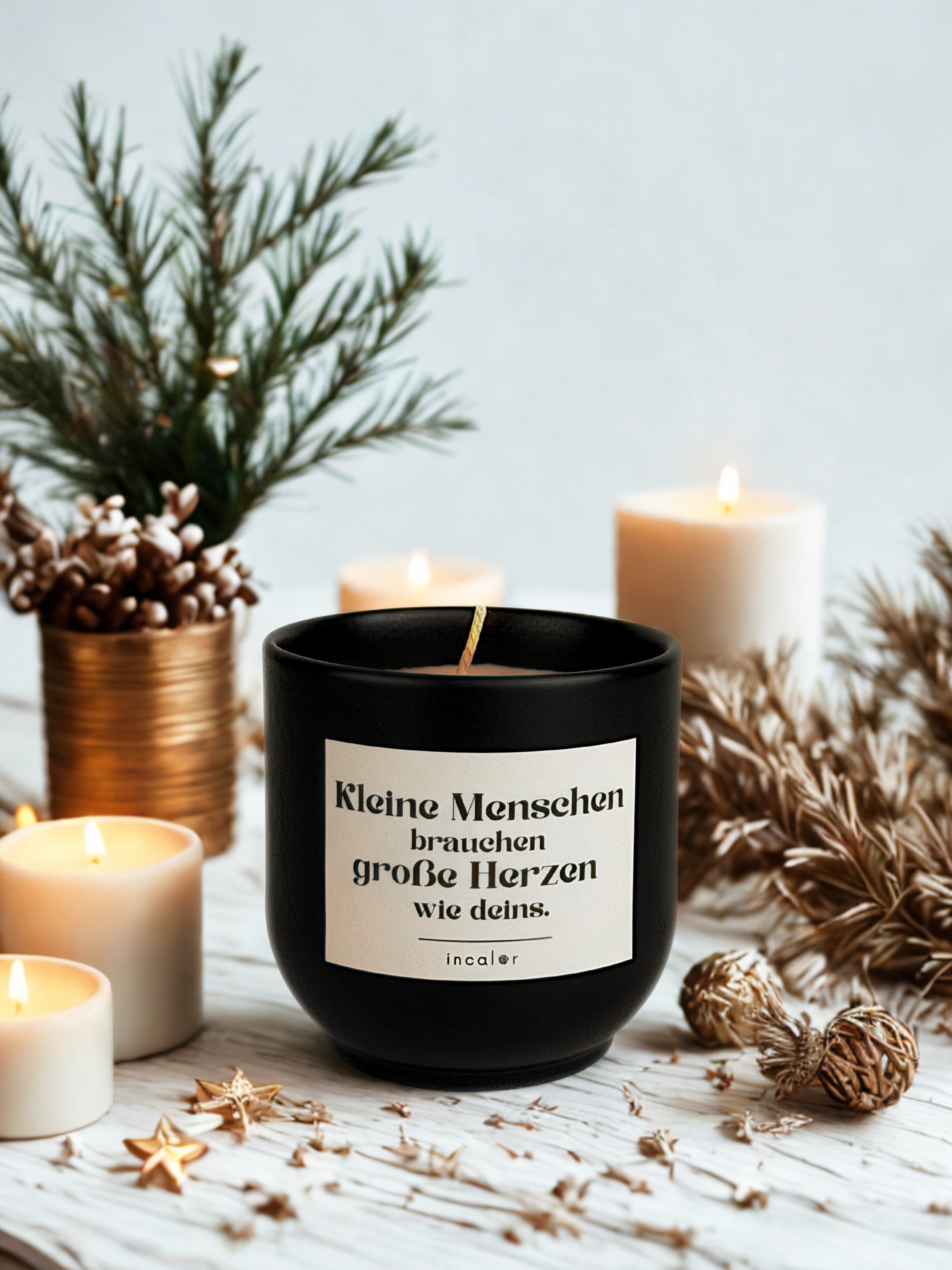 Dunkelgrüne (deep ivy) Duftkerze mit cremefarbenem Etikett und der Aufschrift „Kleine Menschen brauchen große Herzen wie deins.“ steht auf einem hellen, winterlich dekorierten Untergrund. Umgeben wird sie von brennenden weißen Kerzen, Tannenzweigen, goldenen Akzenten und kleinen Sternornamenten, die eine warme, festliche Stimmung erzeugen.