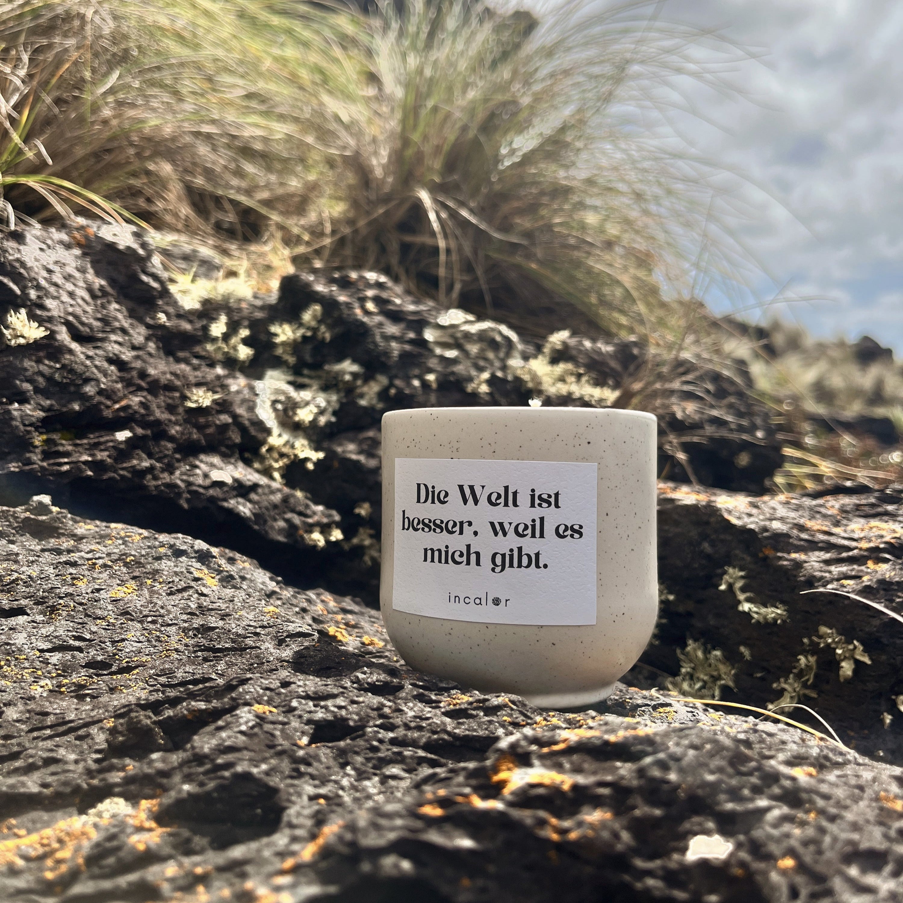 Beige Kerze aus Keramik (Farbton Sahara) mit Affirmation „Die Welt ist besser, weil es mich gibt.“ von incalor, eingebettet in natürliche Vulkanlandschaft mit trockenen Gräsern und Lavagestein – eine Botschaft für Selbstwert und Verbindung zur Natur.