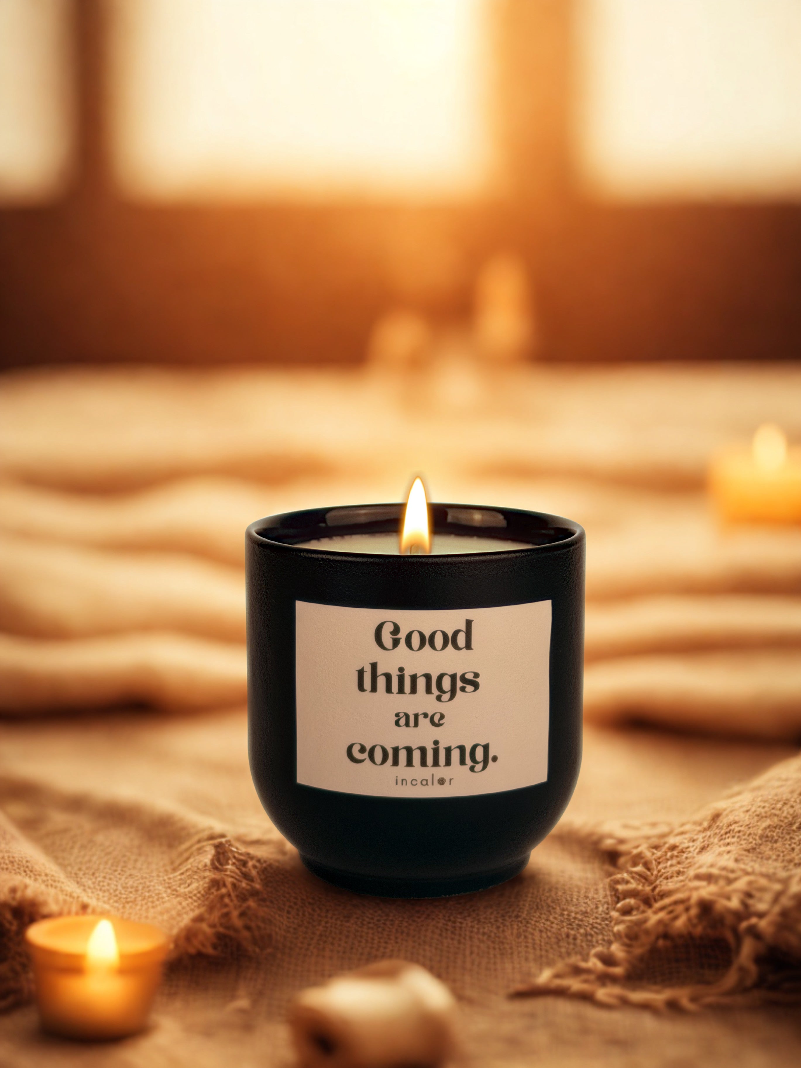 Dunkelgrüne (deep ivy) Duftkerze mit brennender Flamme steht auf einem weichen, beigefarbenen Stoff. Auf dem Etikett steht „Good things are coming.“ und darunter der Markenname „incalor“. Um die Kerze herum liegen kleine Teelichter und Dekorationselemente, während warmes Licht im Hintergrund eine gemütliche Stimmung erzeugt.