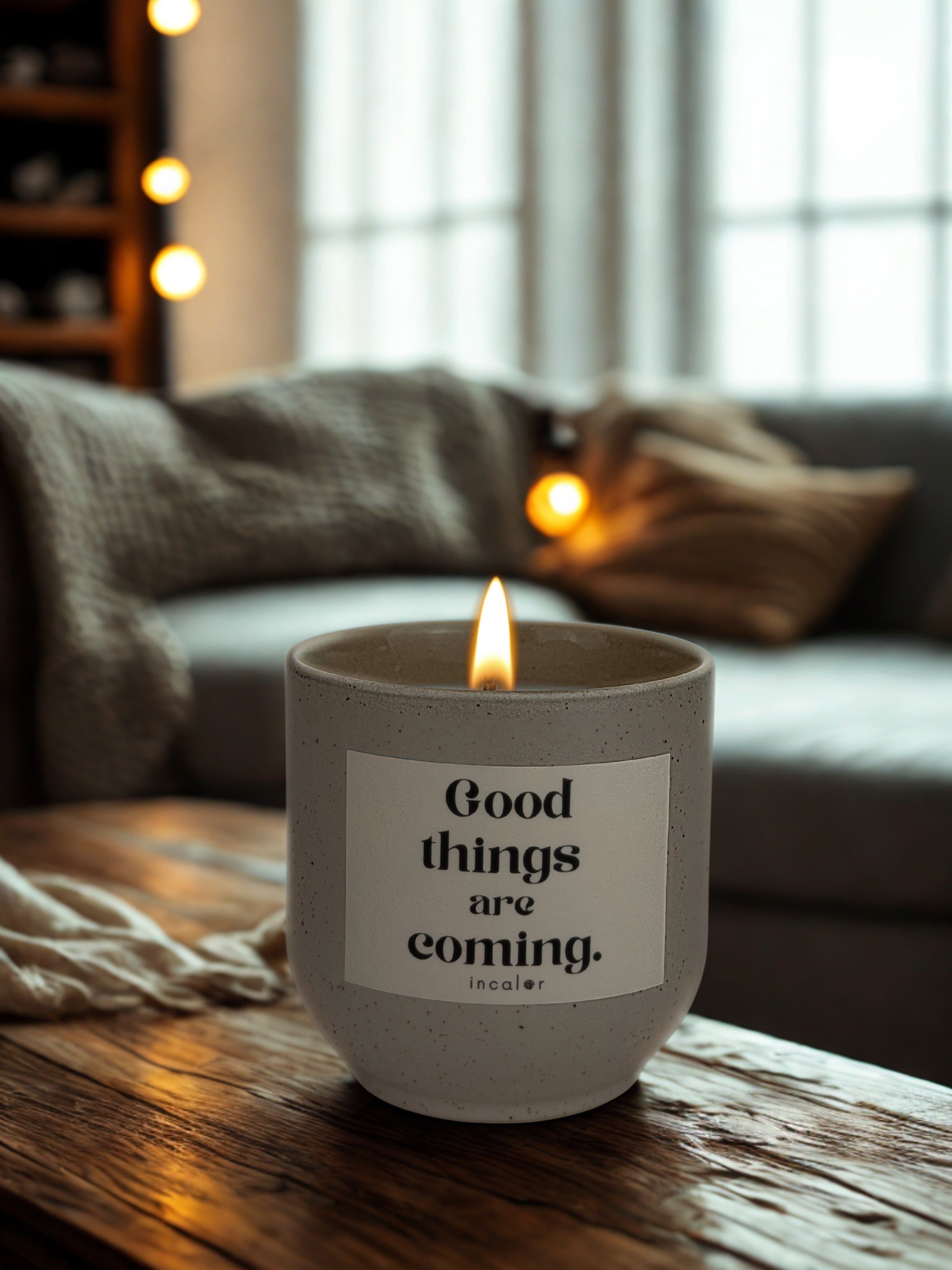 Beigefarbene (sahara) Kerze mit der Aufschrift „Good things are coming.“ von incalor, brennend auf einem Holztisch in einem warmen, gemütlich beleuchteten Wohnzimmer mit Sofa, Kissen und Lichterkette im Hintergrund.