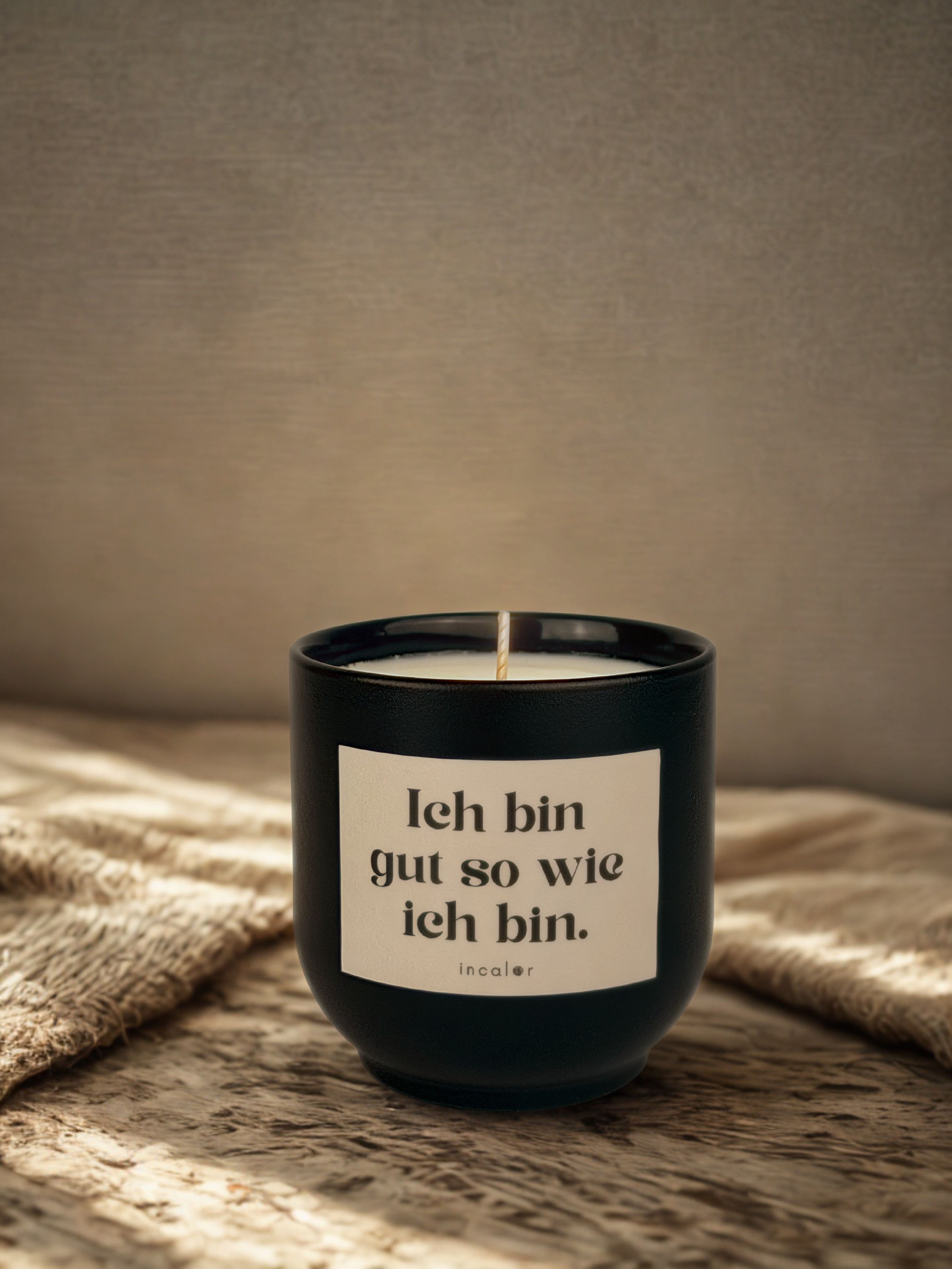 Dunkelgrüne (deep ivy) Duftkerze mit weißem Etikett und der Aufschrift „Ich bin gut so wie ich bin.“ steht auf einer strukturierten Stoffdecke. Warmes Licht fällt von der Seite ein und erzeugt eine ruhige, wohnliche Atmosphäre vor einem sanft strukturierten Hintergrund.